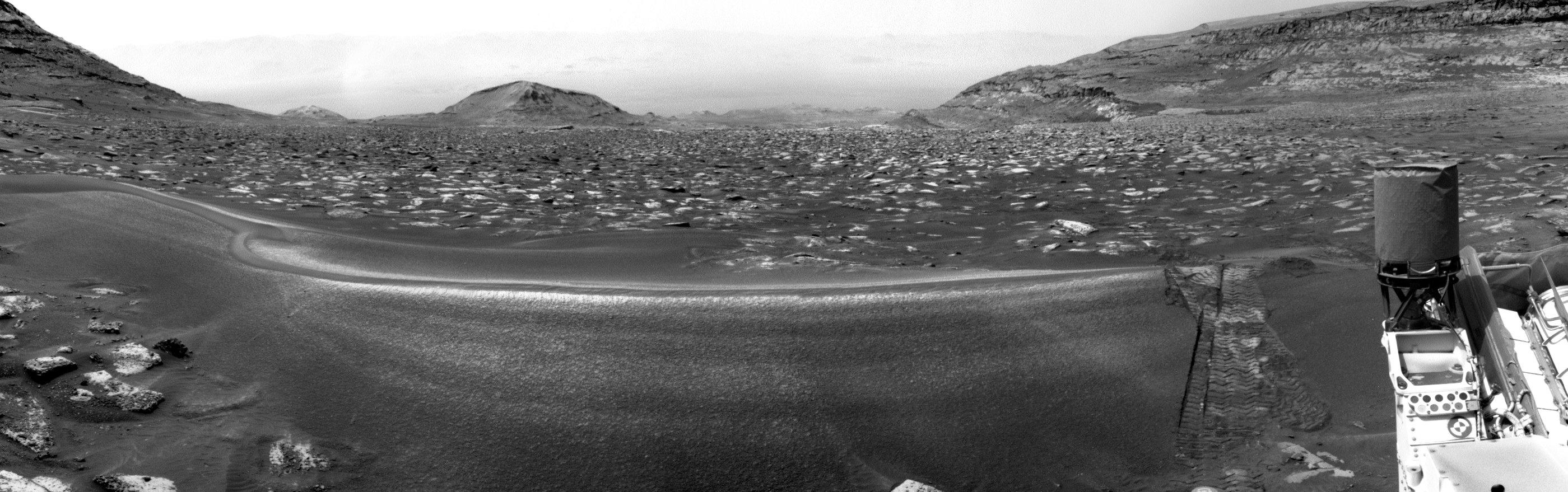 A black and white panorama capturing a Martian landscape, with the UHF antenna of the rover in the foreground to the right. The tracks of the rover go across a fine sand ripple barring the view. This panorama is obtained by stitching together four images acquired by the left navigation camera.