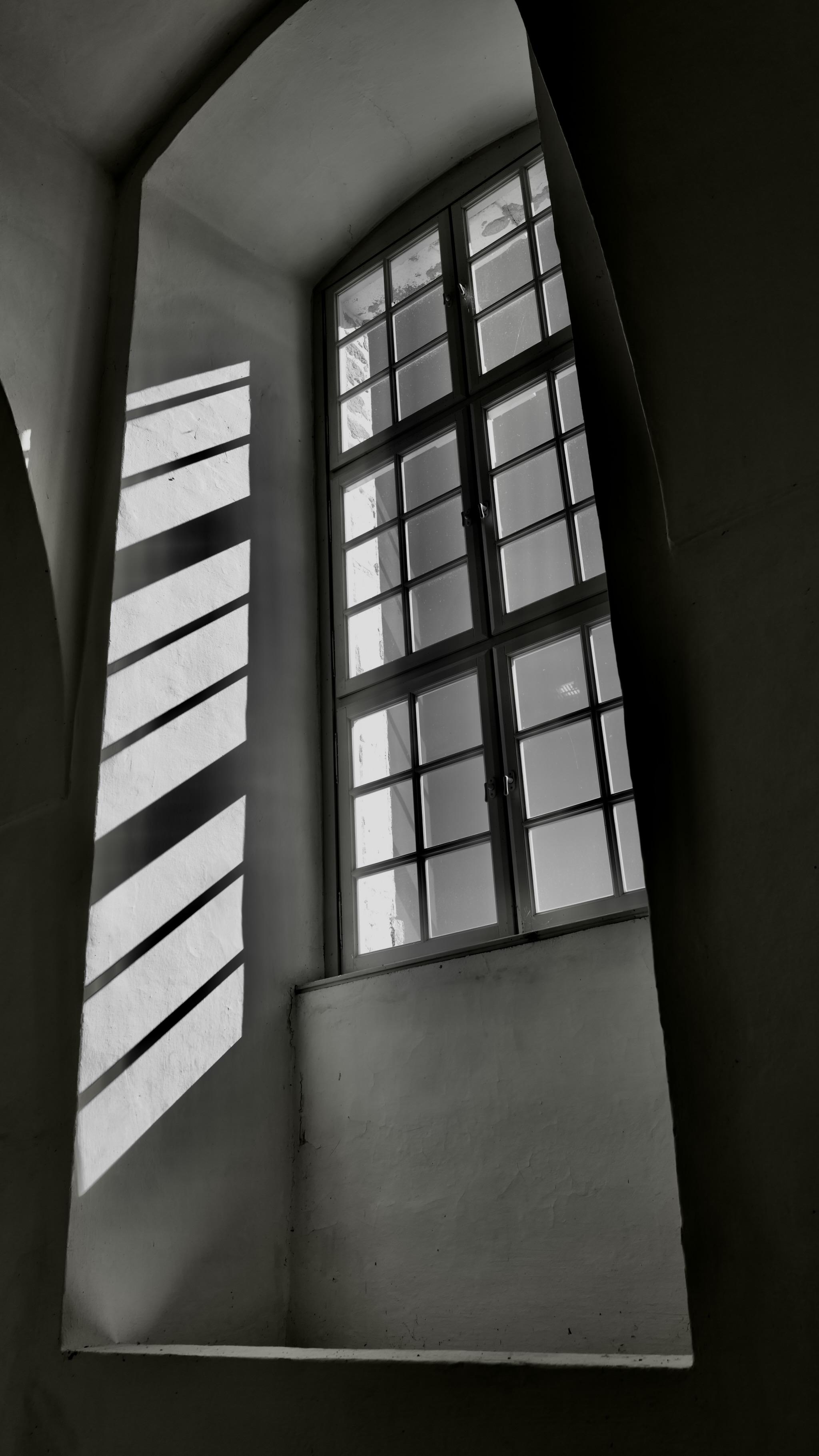 Picture of a window in the Altenburg monastery in Austria. It is located in a high room in a historic building. Sun is casting a bright area inside with a grid of hard shadows on the wall. 
Photo is black and white.