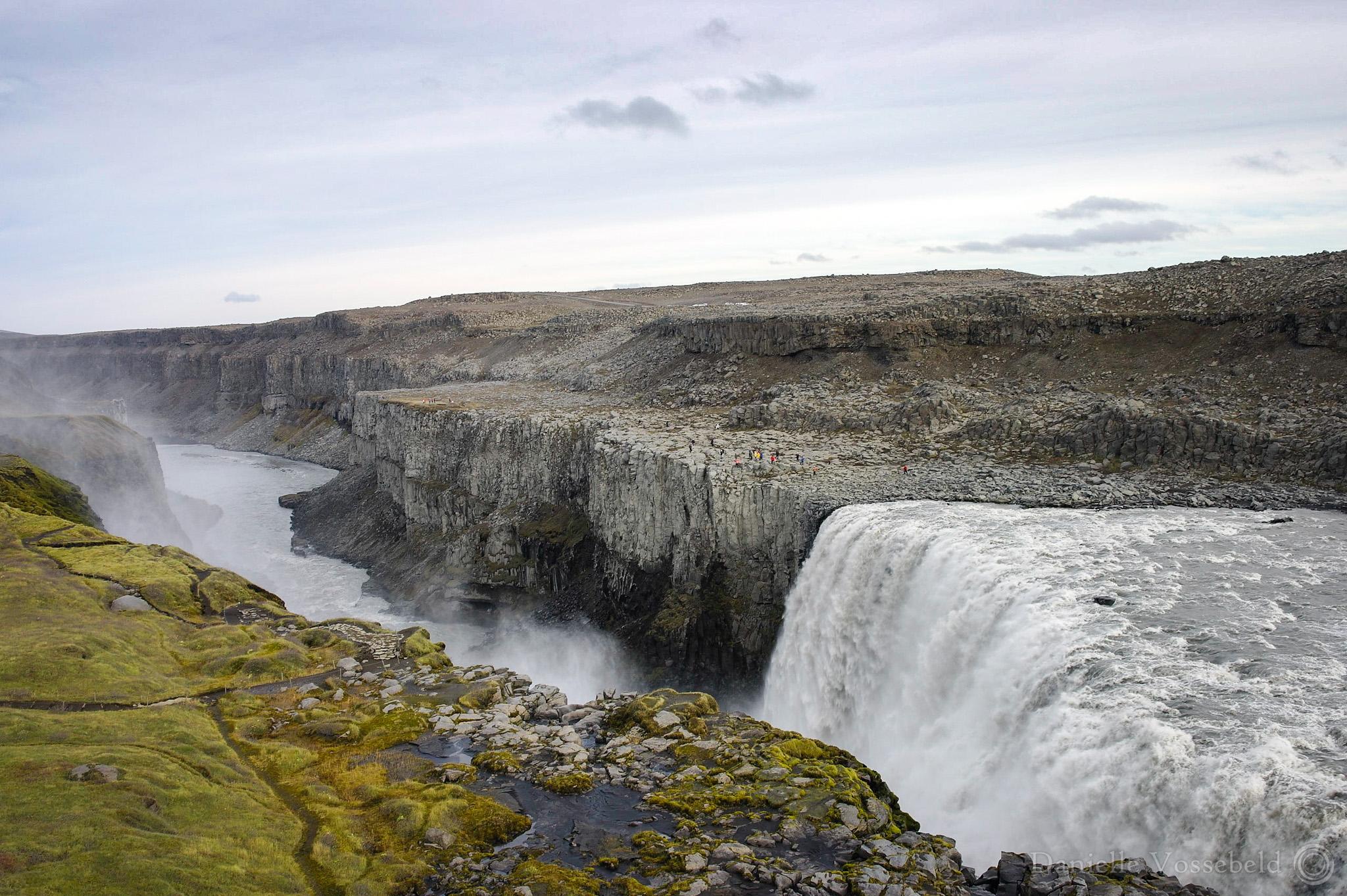 Wide waterfall on the right, on the left of the photo you can see the river continuering. The other side is grey rocks, with tiny colour specks (people). This side is mossy, grass, with some walking paths. The water is water.
