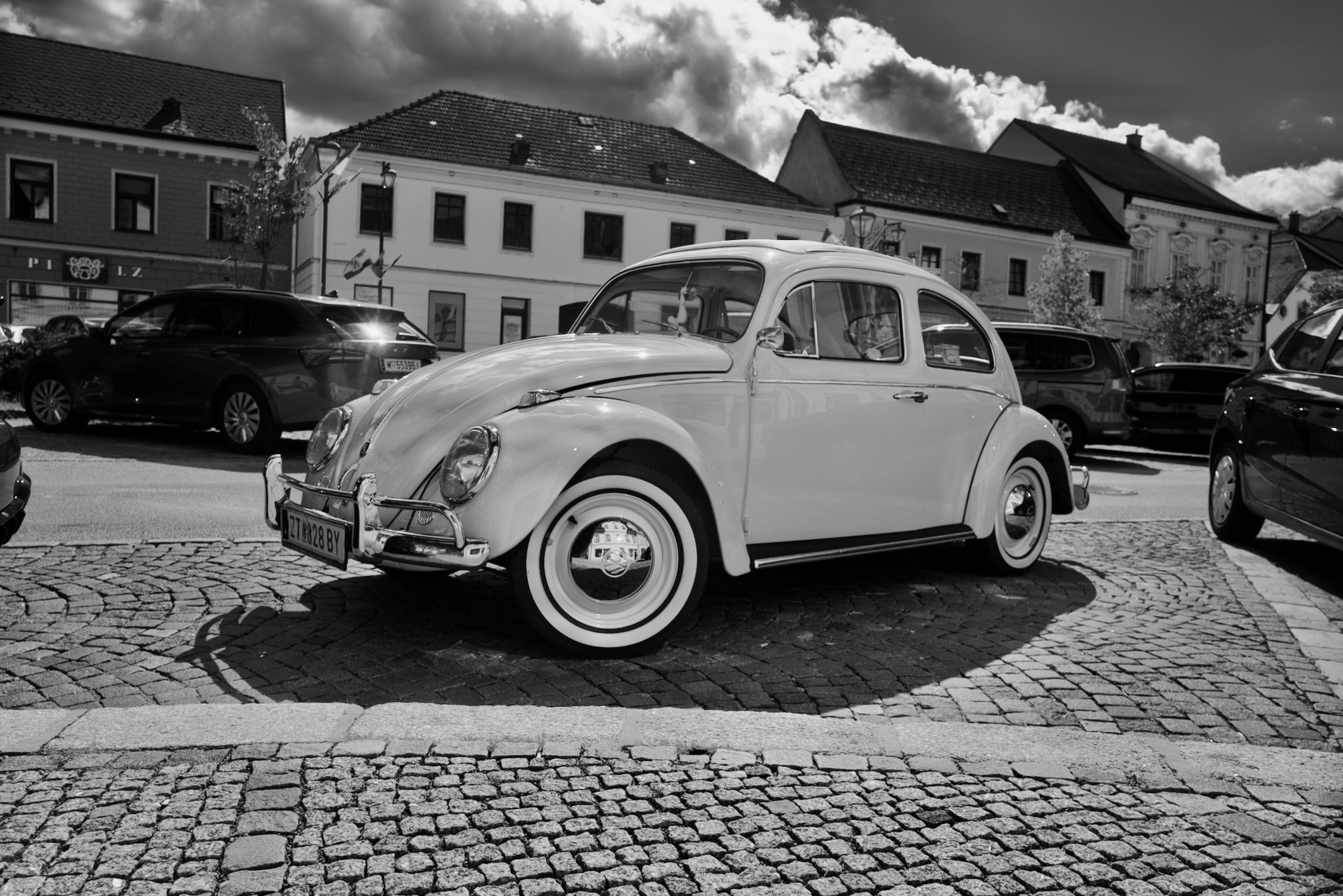 Picture of a VW Beetle on a parking spot in Austria. The shot it taken from the front, showing the drivers side. The car is bright (colour was egg shell), fully in sunlight and shiny with chrome applications. The floor is plaster stones, the background are historical houses on a town square. Some modern cars around are mainly dark and contrasting the oldtimer.
Photo is black and white.