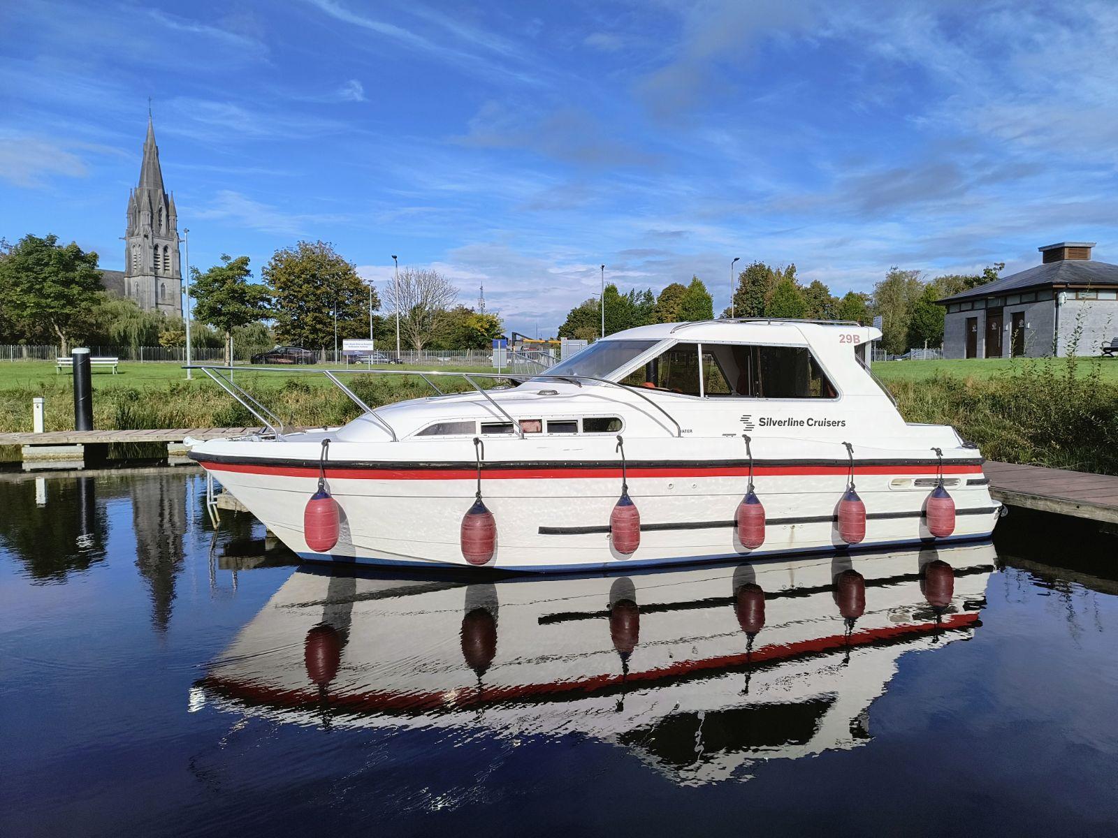 Im Wasser des Hafenbeckens spiegelt sich ein kleines, weißes, modernes Hausboot mit 6 roten Pfendern im Sonnenschein.
Im Hintergrund sieht man das Versorgungsgebäude und den Steinkirchturm von Ballinasloe.