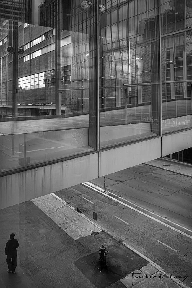 A person seen walking the street, near the lower edge of a tall building with multiple reflections.
