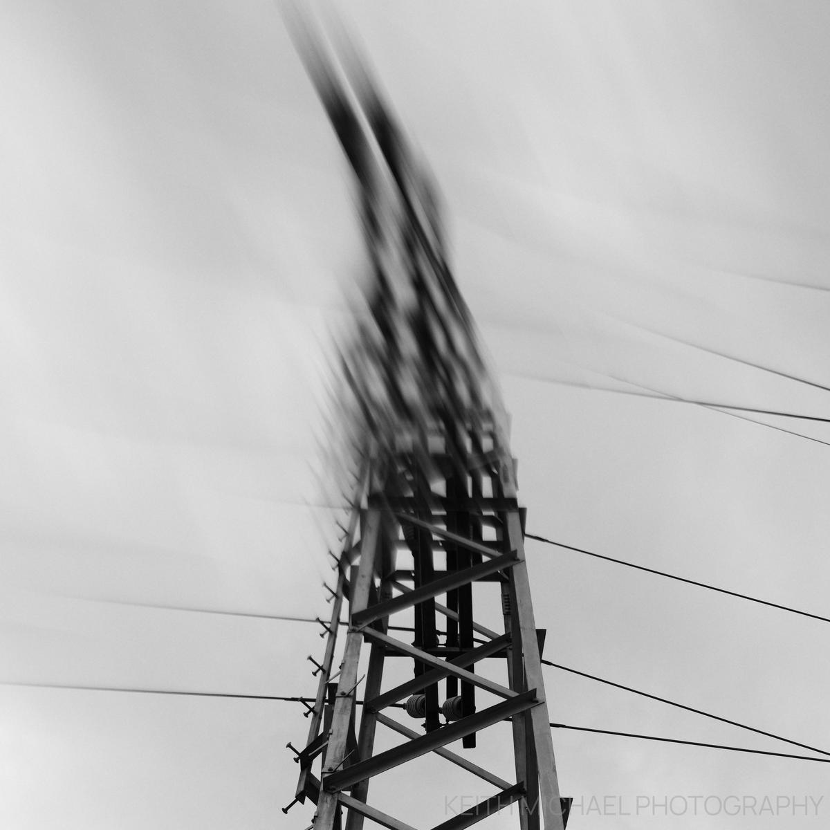 A monochrome photograph of a powerline tower that is blurred from the top half off to the left.