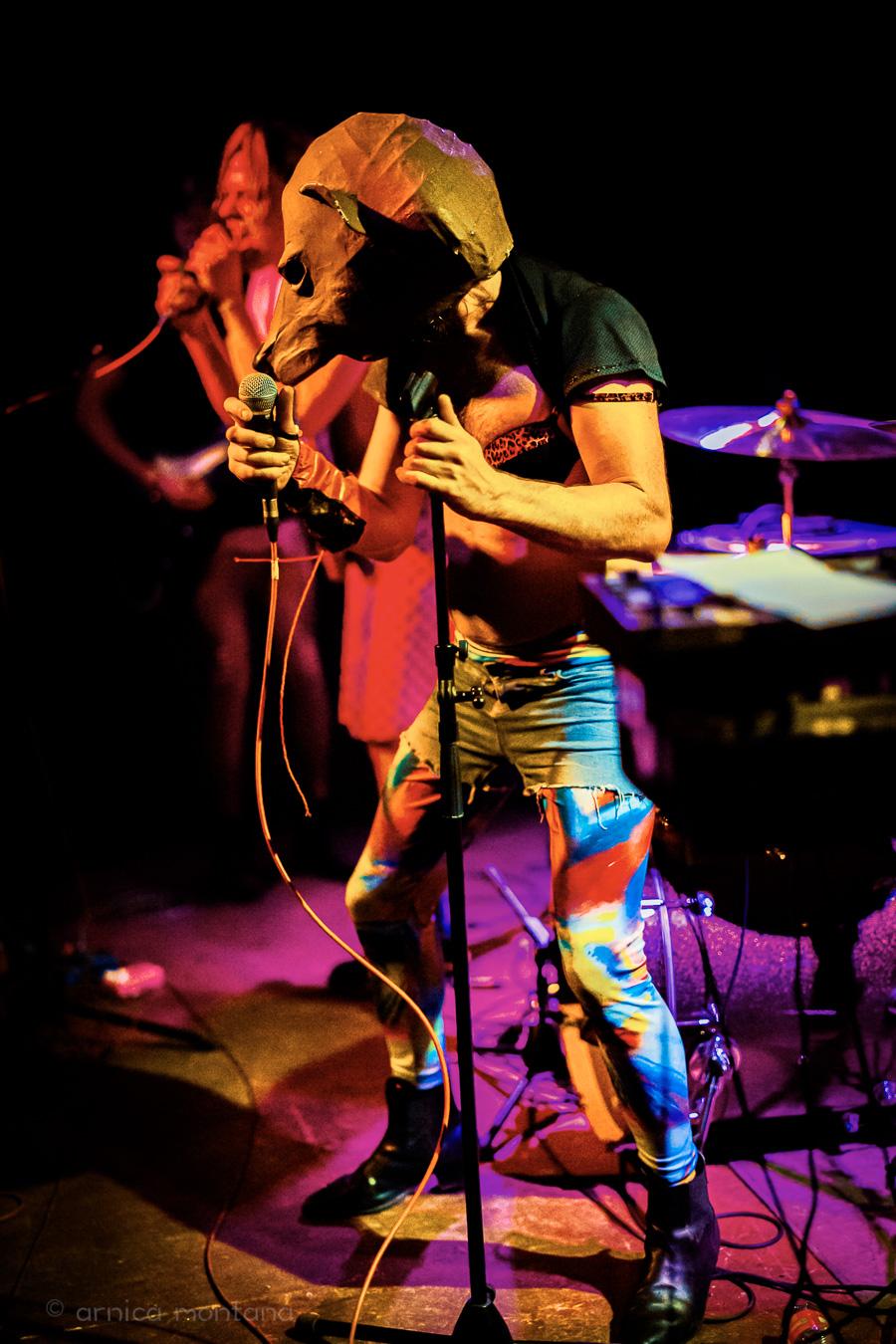 A person wearing an animal mask is singing into a microphone. They are wearing brightly colored leggings, denim shorts, and a leopard-print bra. In the background, under the stage lights, is a second singer and a keyboard.