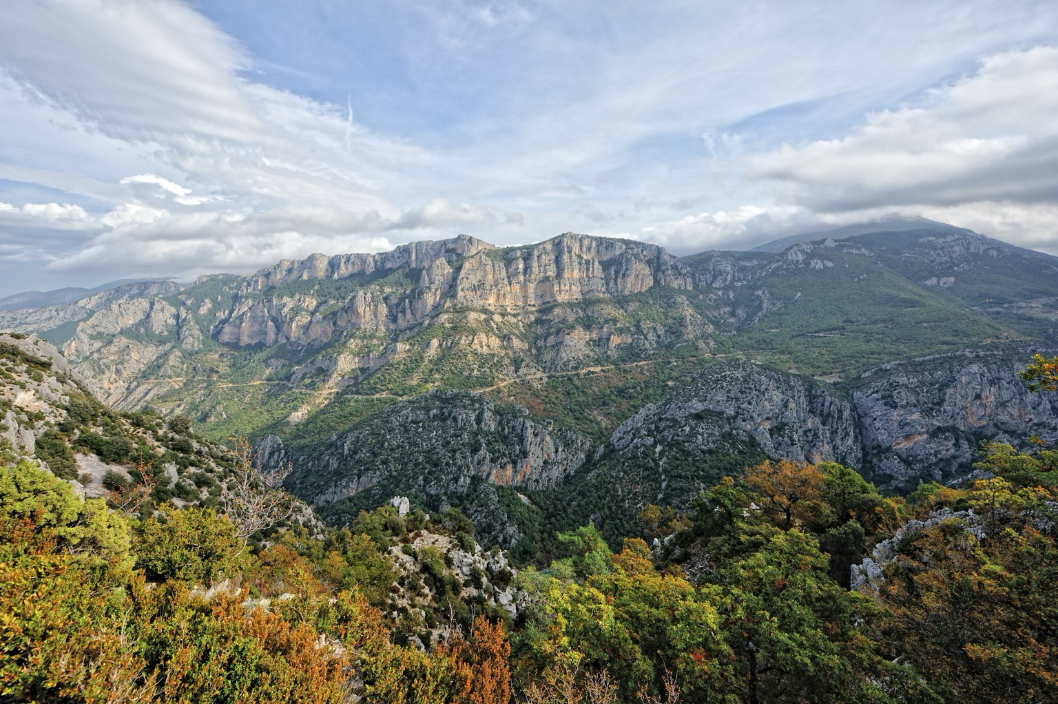 Eine weite Gebirgslandschaft. Im Vordergrund befinden sich Büsche und Bäume mit grünen, gelben und orangefarbenen Blättern. Dahinter erstreckt sich ein tiefes, dicht bewaldetes Tal mit felsigen Einschlüssen. Im mittleren Hintergrund erhebt sich ein markantes Bergmassiv mit steilen, hellen Felswänden, die einen langen Kamm bilden. Weitere, sanftere Berge verblassen am Horizont. Der Himmel ist von ausgedehnten weißen und grauen Wolkenformationen mit dazwischenliegenden blauen Flächen bedeckt. A vast mountainous landscape. In the foreground, there are bushes and trees with green, yellow, and orange leaves. Behind this extends a deep, densely wooded valley with rocky inclusions. In the middle background rises a prominent mountain range with steep, light-colored rock faces forming a long ridge. Other, gentler mountains fade on the horizon. The sky is covered by extensive white and gray cloud formations with interspersed patches of blue.