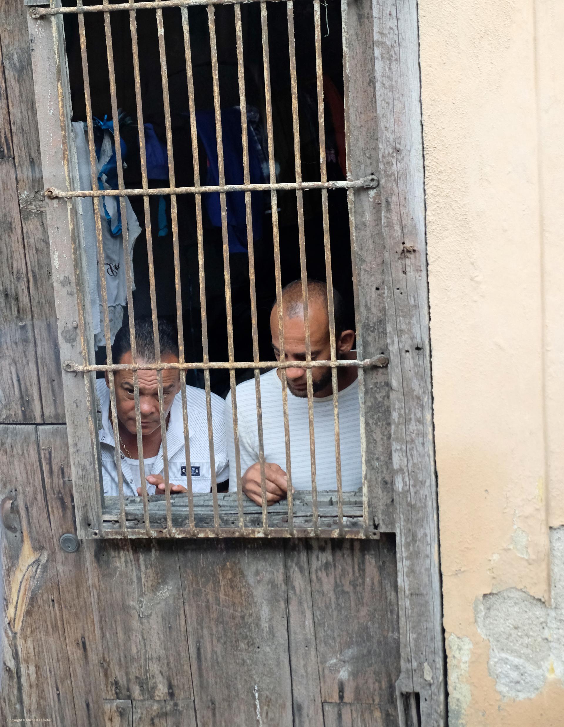 Photo by Fenichel: "Looking Out (Cienfuegos, Cuba)"

Two men peer out from behind a barred window, framed by an old wooden wall and crumbling concrete.