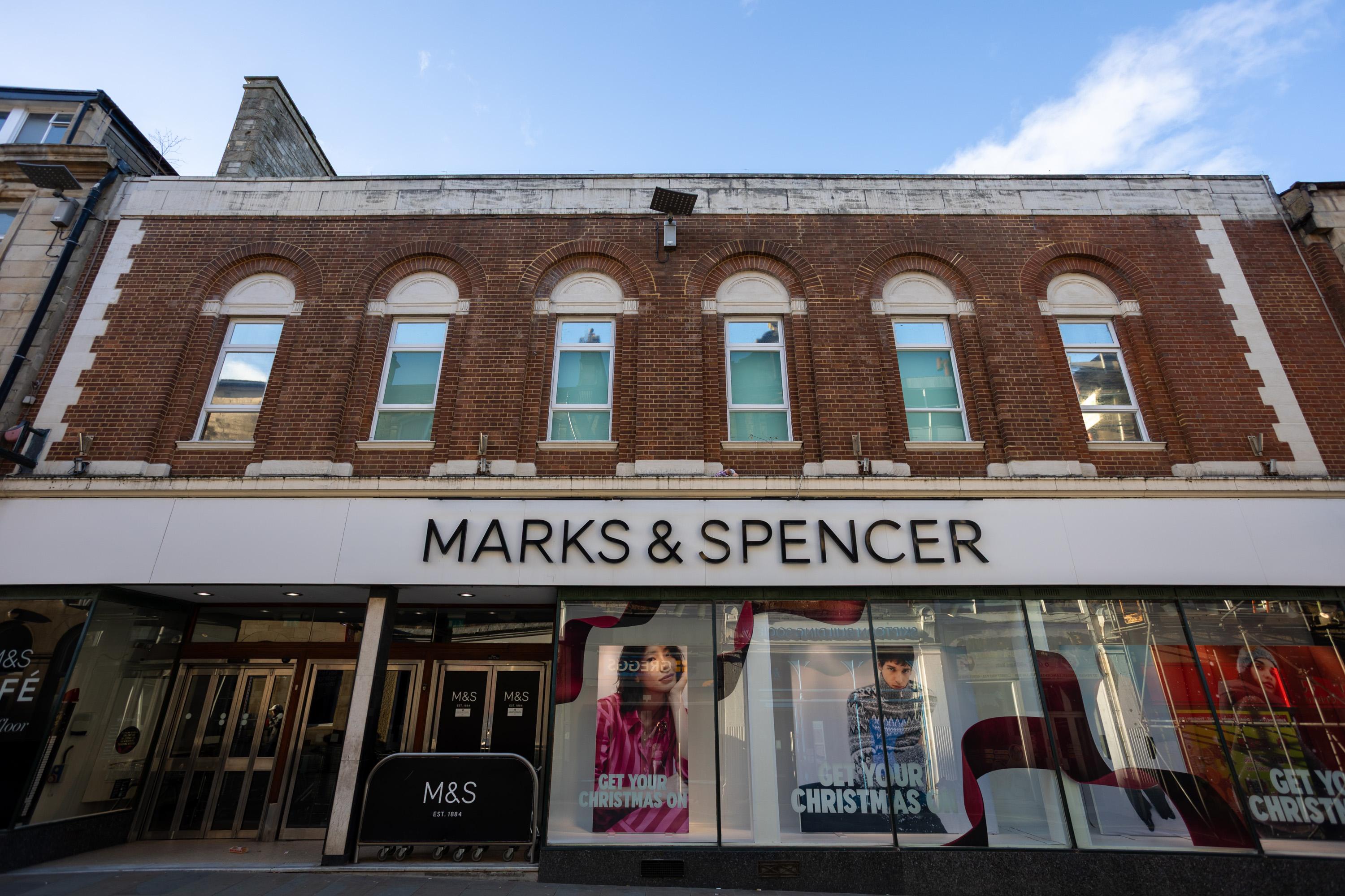 A Marks & Spencer shopfront with a brown brick façade, arched upper windows, and large display windows below featuring Christmas-themed posters. The white sign above the entrance displays the shop name in bold black letters. Blue sky is visible above.