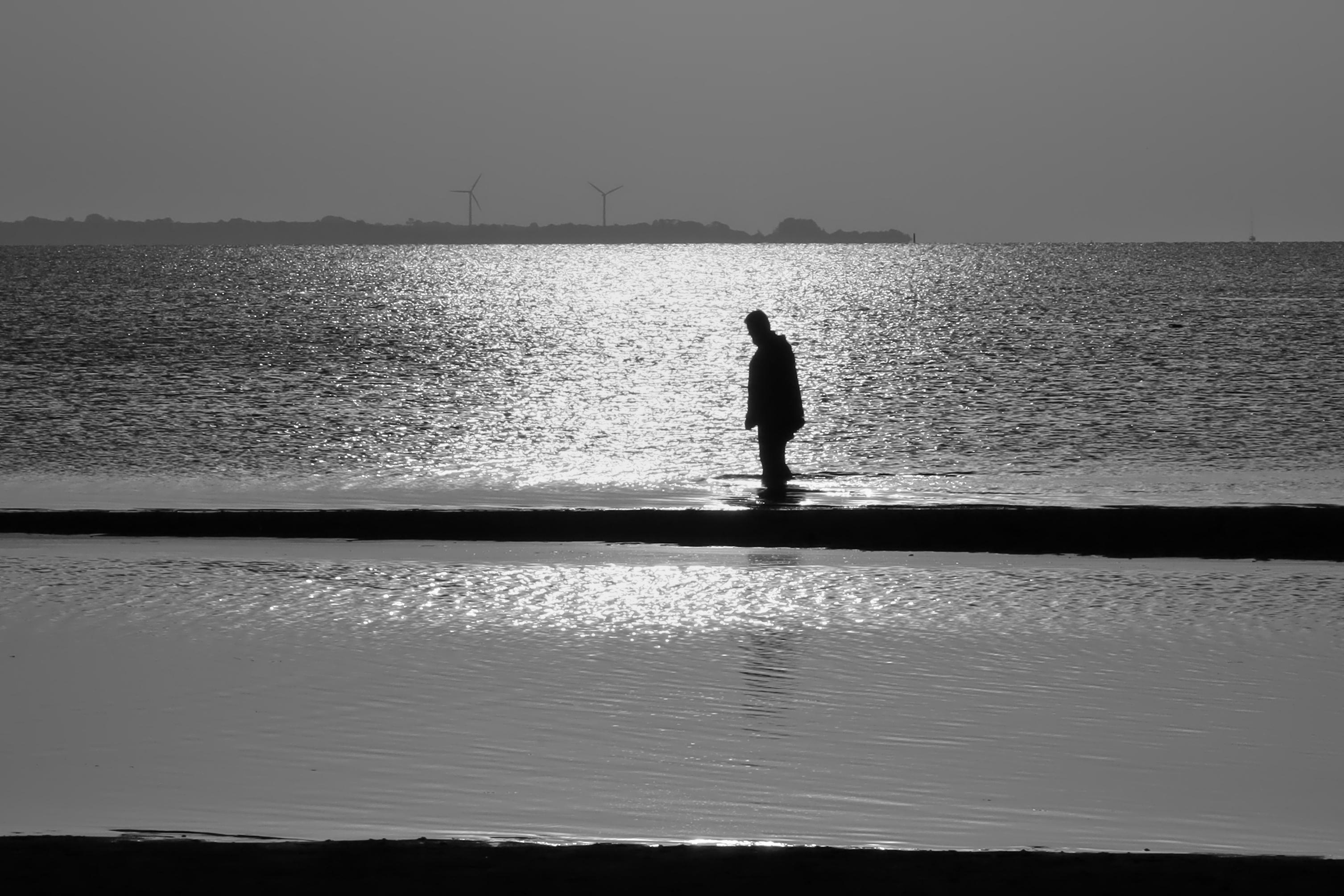 Gegenlicht-Aufnahme in Schwarzweiss. Gezeigt wird ein Sonnenaufgang an der runigen Ostsee vor der Halbinsel Holnis an der dänischen Grenze. Das Sonnenlicht spiegelt sich auf den Wasser und lässt den morgendlichen Wanderer als Silhouette erscheinen.