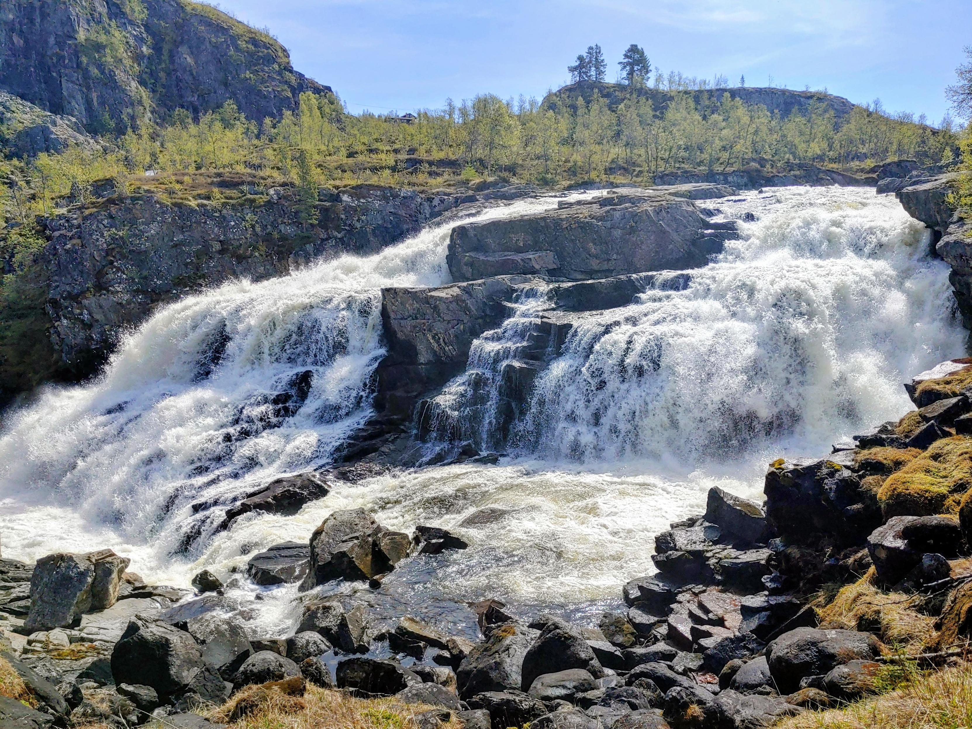 Wasser fließt kaskadenförmig über scharfkantige Felsen umgeben von einer bergigen Landschaft.