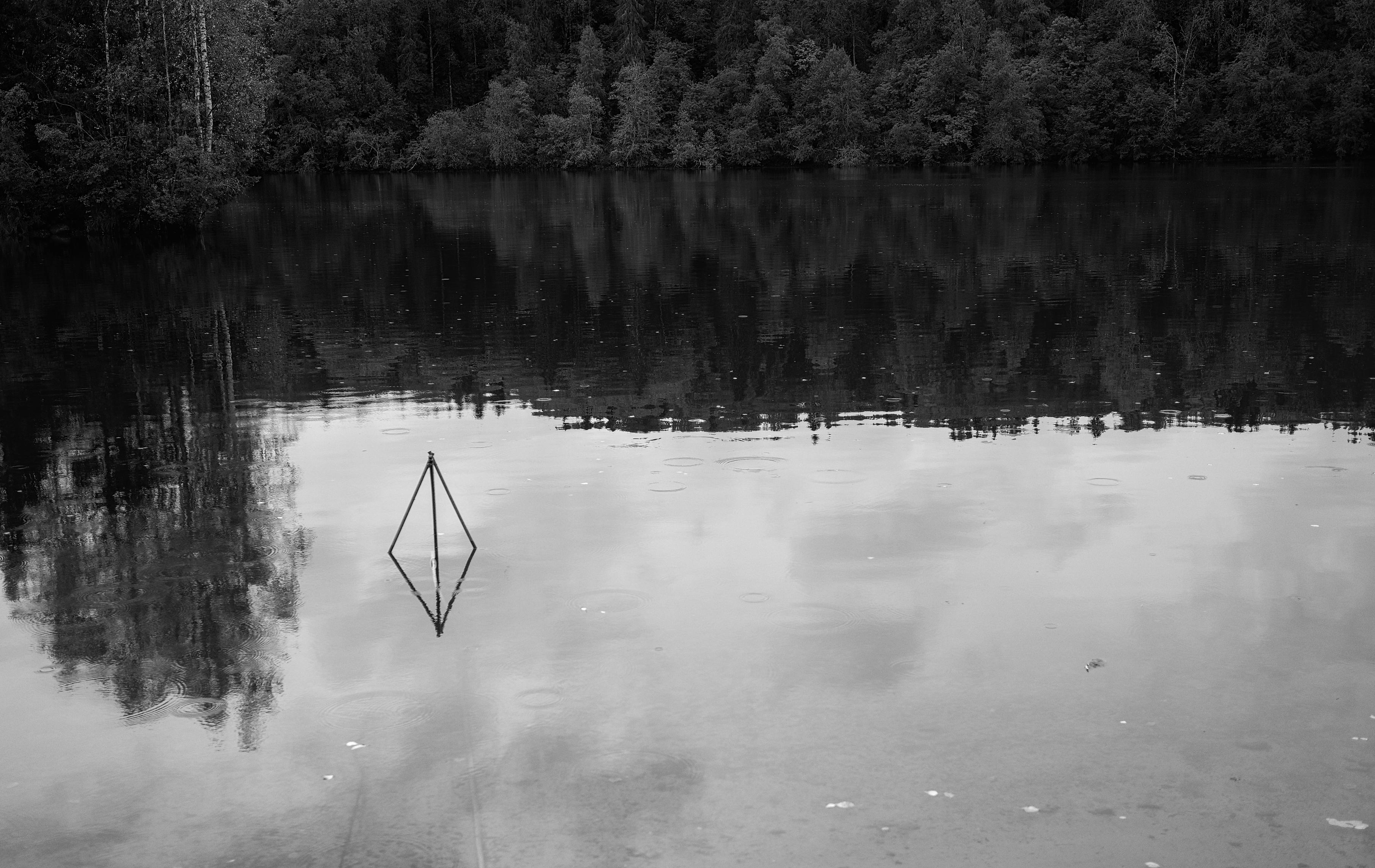 This black-and-white photograph captures the calm surface of water, likely a forest lake. In the upper part of the frame is a dense wall of dark forest, which reflects in the water, creating a massive, opaque patch. The lower and right parts are light, mirror-like water reflecting the cloudy sky. In the center of this boundary, on the water, stands a perfect geometric object — a diamond (likely the frame of a fishing trap). Its clear, light lines sharply contrast with the dark reflections of the forest, creating the effect of a "portal" or a sign. The image conveys a sense of absolute silence, solitude, and mysterious harmony between geometry and wild nature.