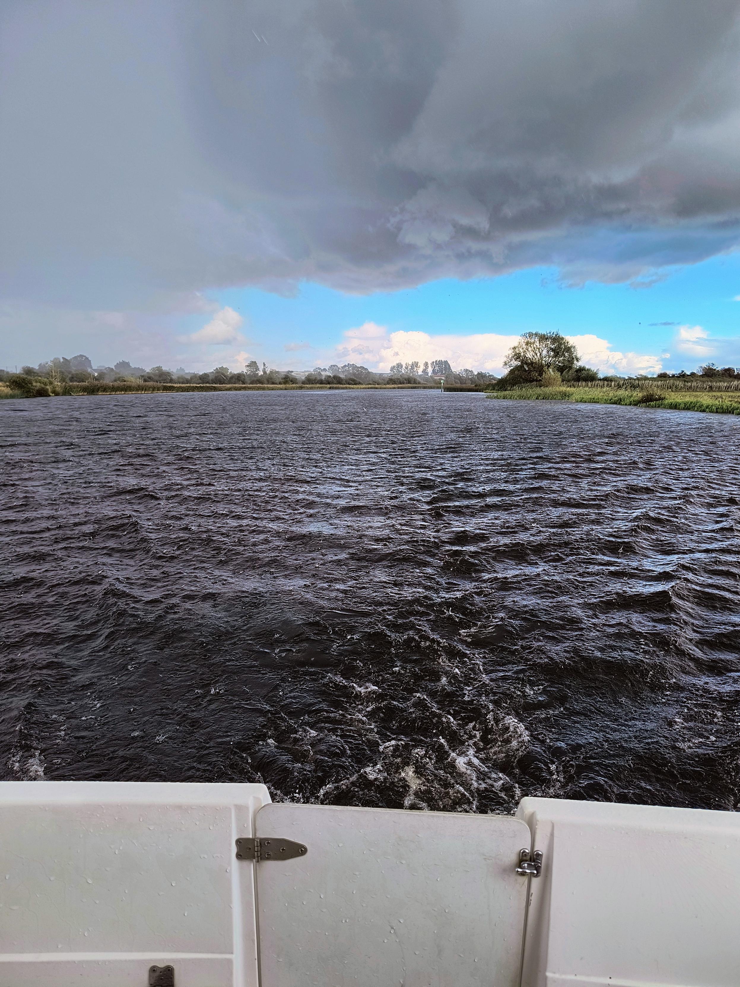 Von der Rückseite eines Schiffes, welches mittig auf dem Fluss fährt schaut man in das aufgewühlte Wasser. Links am Horizont erkennt man das es regnet, während Rechts noch blauer Himmel und Sonnenschein zu sehen ist. Über allem türmen sich die grauen Regenwolken auf.