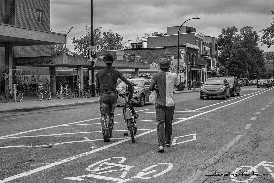 Two men crossing a street alongside one bicycle, One of them wearing a cowboy hat, with attitude.