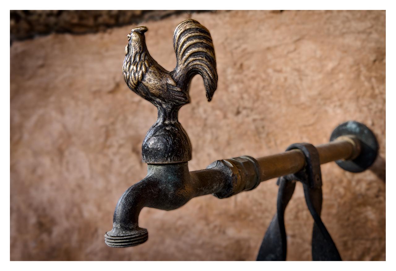 Close-up of an antique brass faucet with a detailed rooster-shaped handle, showing weathered patina and green oxidation, mounted against a textured beige plaster wall with a metal pipe.