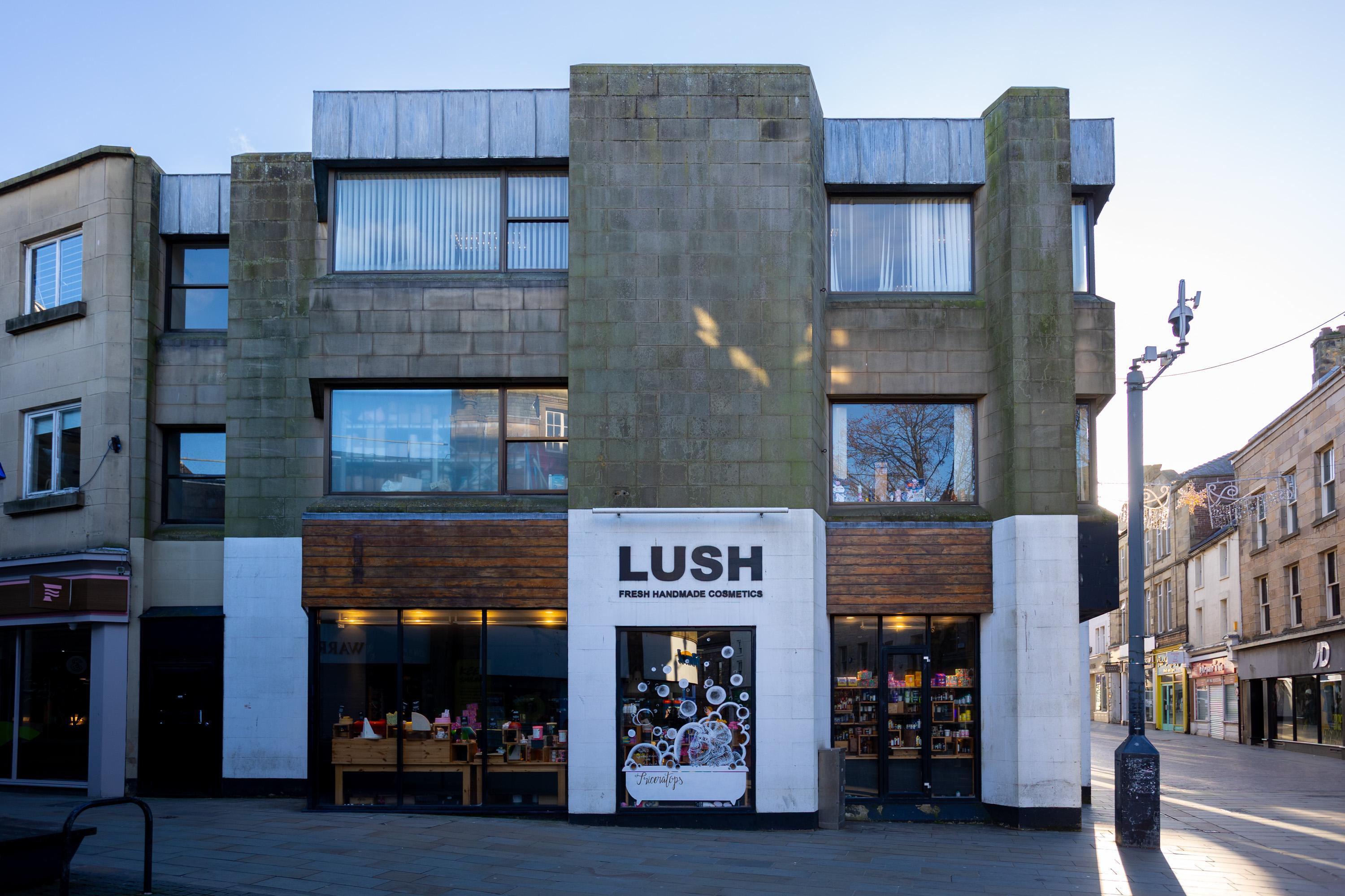 A shopfront for LUSH, a handmade cosmetics shop, is in a modern, rectangular stone building. The large white sign displays LUSH over glass windows showing products. The street in front is empty, and sunlight illuminates the scene.