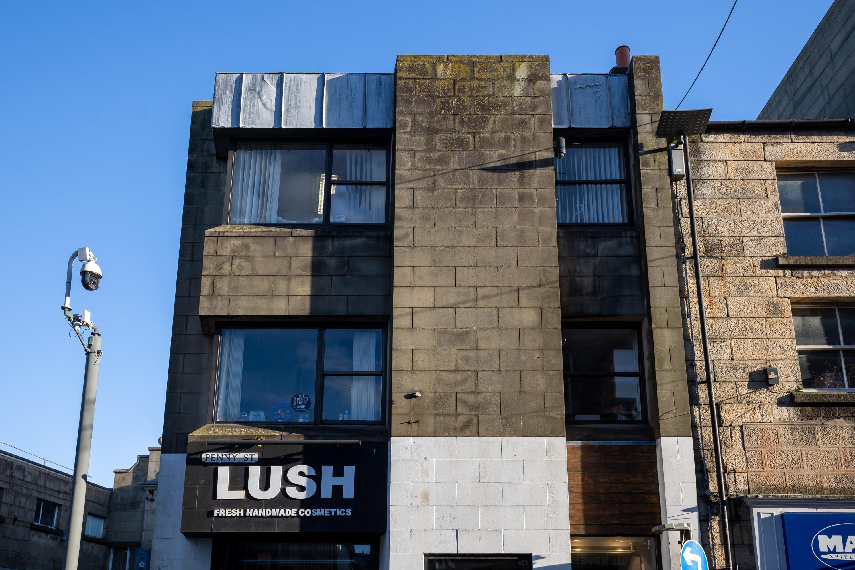 A three-storey stone building with a LUSH Fresh Handmade Cosmetics shop on the ground floor. The upper floors have large, square windows and stained, weathered walls. A CCTV camera is mounted on the left. Bright blue sky overhead.