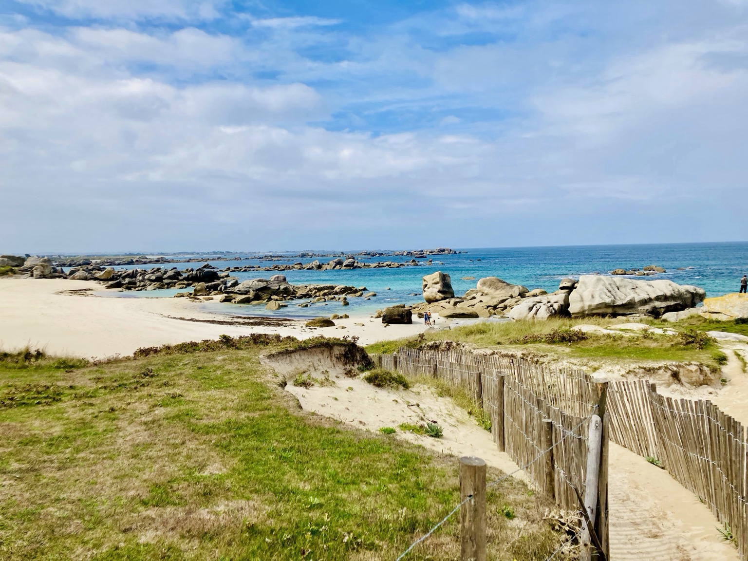 Ménéham. Kerlouan. Plage de sable au bout d’un sentier vert bordé d’une barrière en bois. Rochers de granit parsemés, laissant entrevoir une mer turquoise et peu profonde.