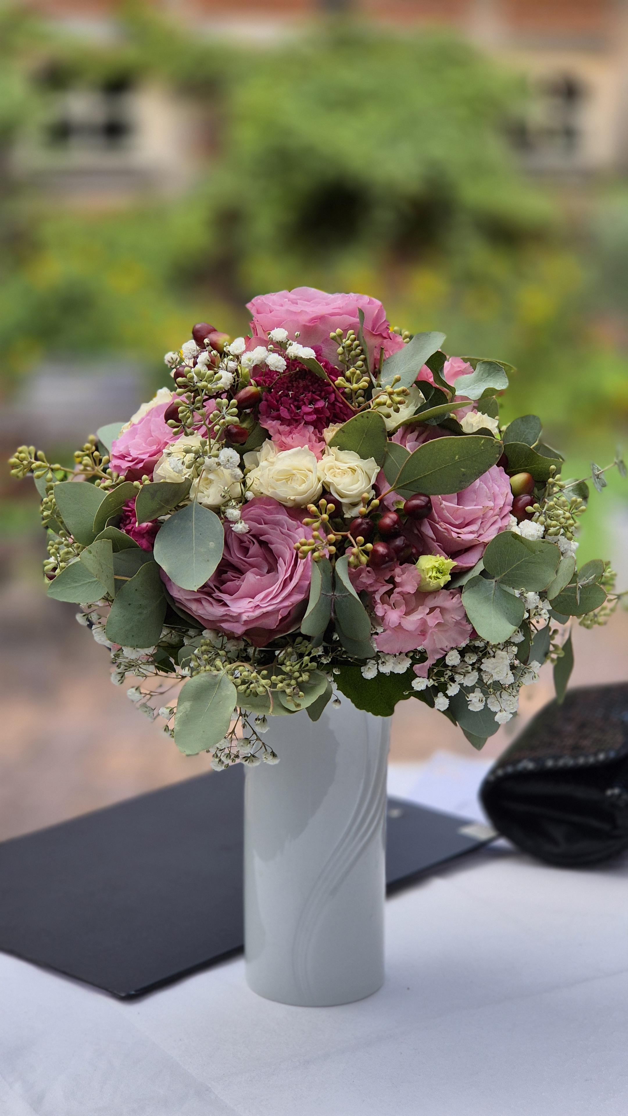 Ein Hochzeitsstrauß mit vielen Rosen in einer weißen Vase auf einem Tischchen vor verschwommenem Hintergrund. /
A wedding bouquet with lots of roses in a white vase on a small table against a blurred background.
