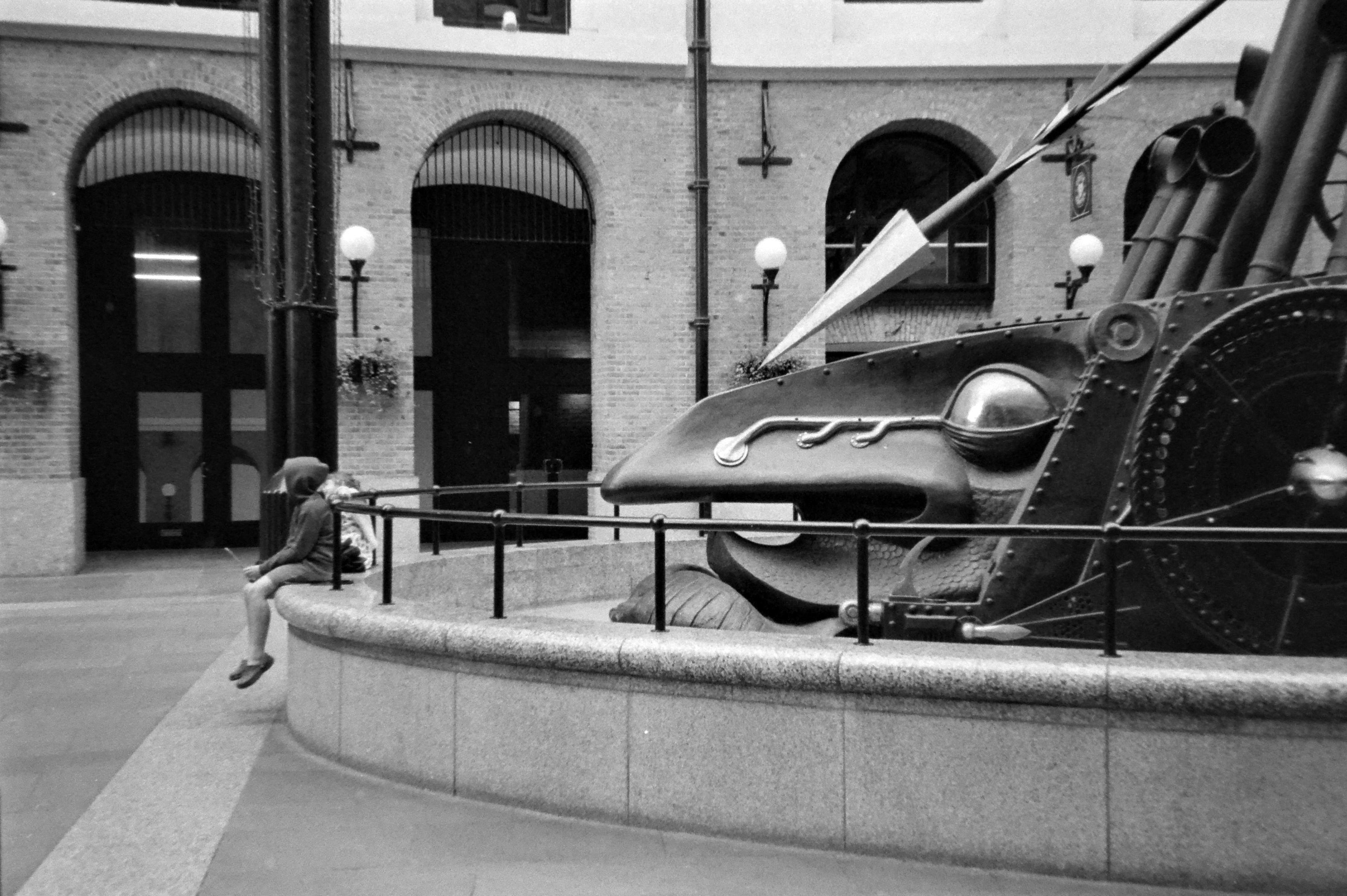 une personne assise au bord d'un bassin dans lequel se trouve une enorme statue métallique qui semble la menacer