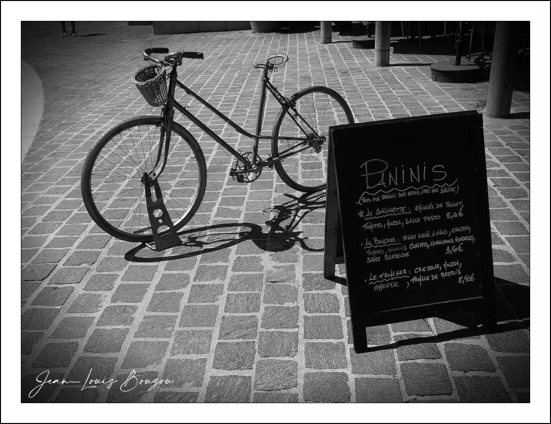 The image is a black and white photograph featuring a bicycle and a sandwich board menu placed on a cobblestone surface. The scene is outdoors, likely in a town square or sidewalk area, suggested by the paved ground and the presence of outdoor furniture or structures in the background, though they are out of focus.
The primary subject is a vintage-style bicycle parked in front of a dark sandwich board. The bicycle is a dark color, possibly black or a dark grey, with a wicker basket mounted on the handlebars. The bicycle is propped upright by a metal stand, which is an interesting design with circular cutouts. The front wheel of the bicycle is large and thin-spoked, and the rear wheel, while less visible, also appears typical for its era. The bicycle casts a distinct shadow on the cobblestones, indicating a strong light source, likely the sun, from the left side of the frame.
The sandwich board is black and has a menu written in white chalk. The word "PANINIS" is prominently displayed a