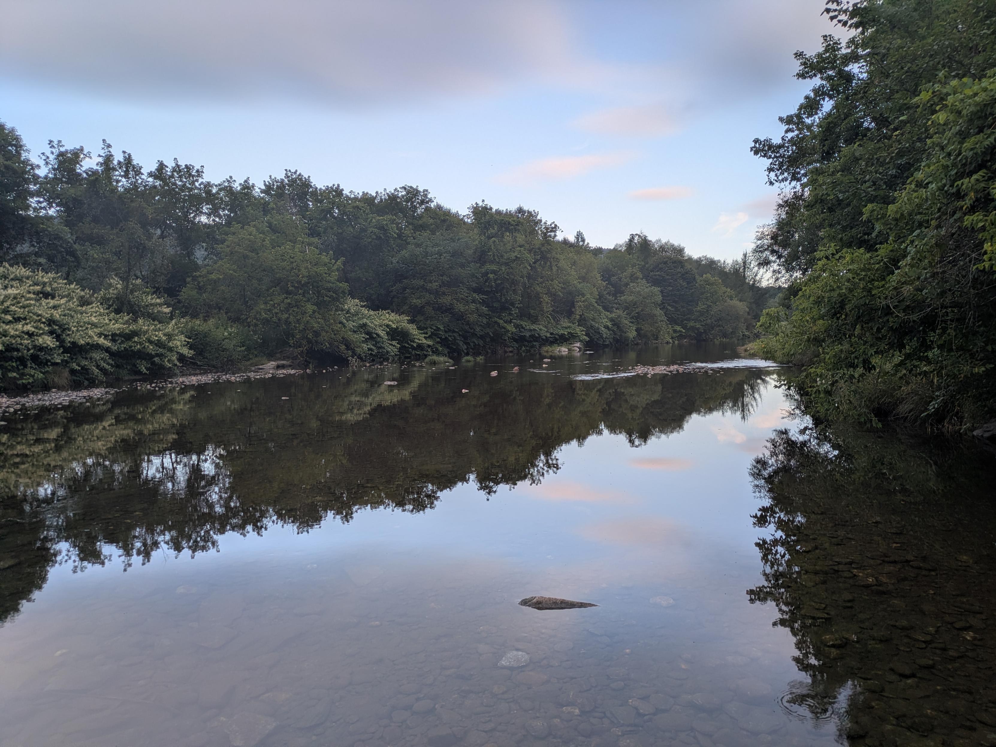 Photo of an early morning landscape with a calm river reflecting tree-lined banks under a partly cloudy sky. The water is so still that it looks like a mirror.