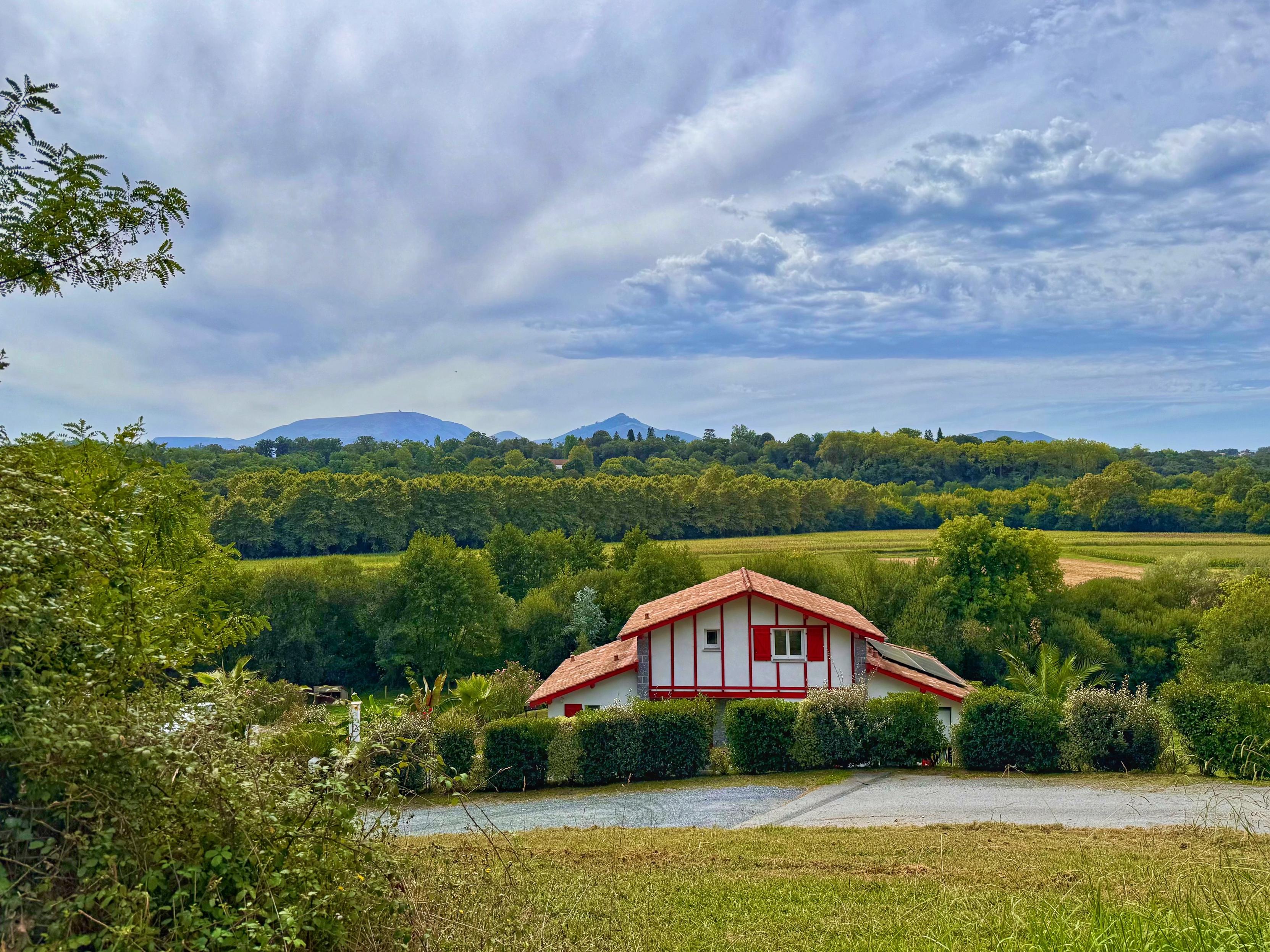 A panoramic scene featuring a somewhat typical Basque house with red timbers, shutters and white walls, surrounded by greenery and trees. Rolling hills are visible in the background under a cloudy sky. Two mountains emerge from the green horizon, one rounded and the other one pointed.