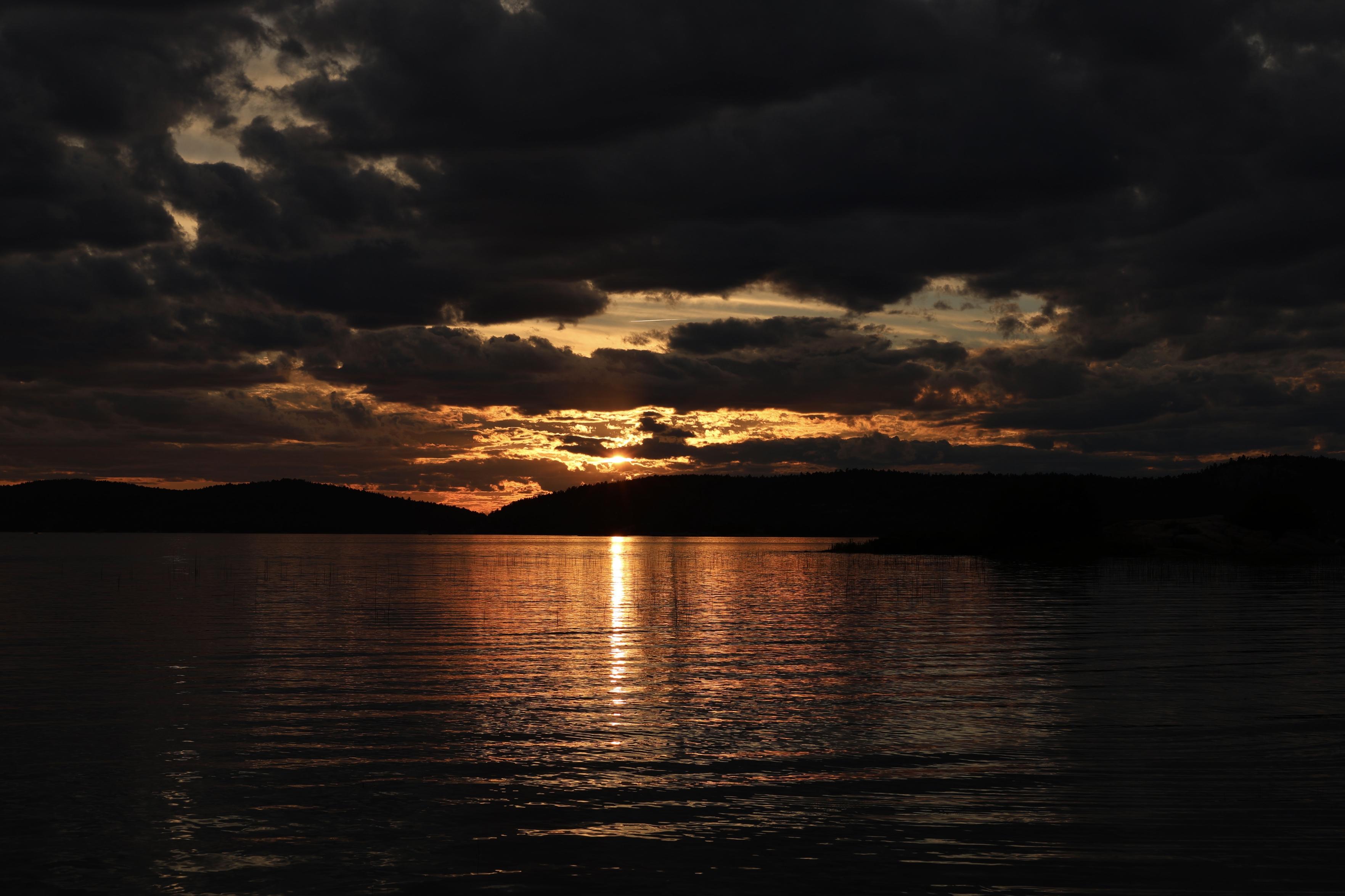 This is landscape format photo of a sunset over a larger lake in the summer. The sky is mostly filled with clouds, some of which are partially shrouding the sun. The warm glow is reflecting on the lake water, as small waves come into shore.
