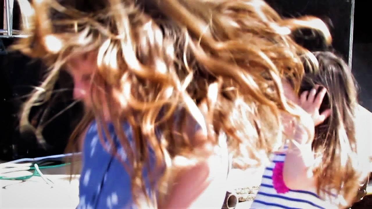 Close up photo of 2 little girls on the grass in front of a live music stage in a park on a sunny day. The girl in the foreground has a blue dress and long golden hair. She is absorbed in the music and dancing so enthusiastically that her bouncing tresses of hair catch the sunlight and dominate the photo. Behind her the 2nd girl, with brown hair and a white and blue stripey top with pink cuffs, is looking up at the band and and clasping her hands to her ears.