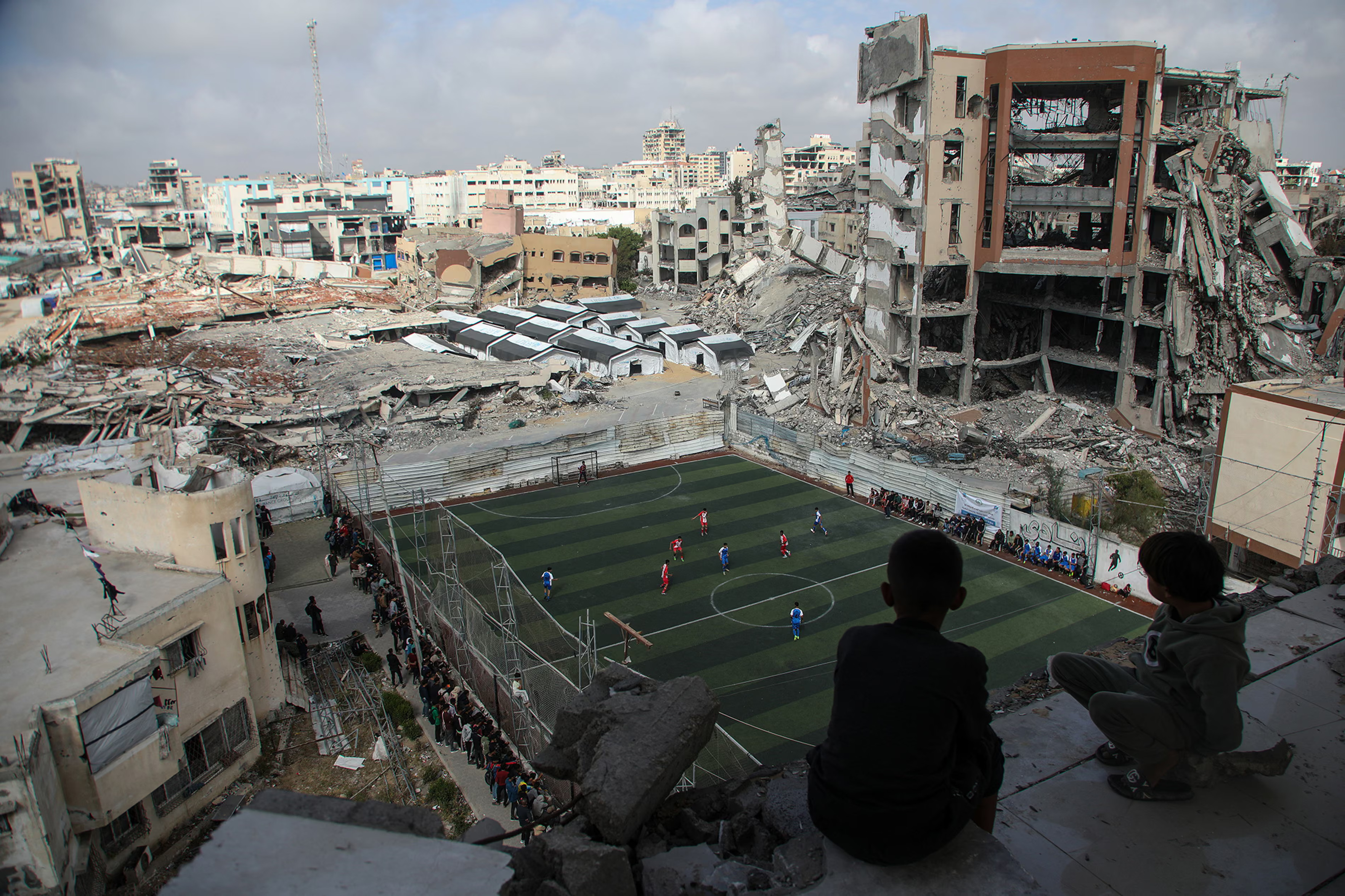 Children watch a football match among the ruins.