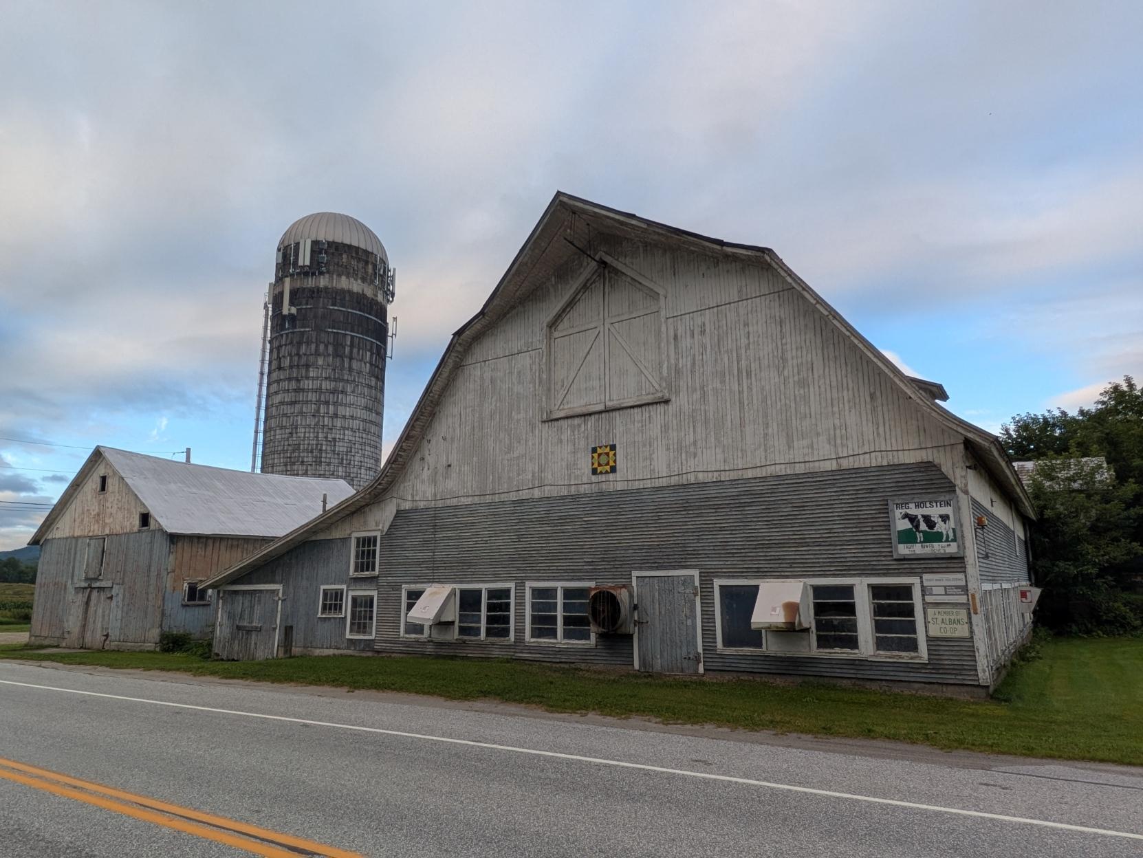 Photo of a white and gray wooden barn, the similarly colored shed next to it and a single large white and gray silo behind that. The signs described in another photo in this post are on the right edge of the barn. The photo is taken from across a light gray blacktop road with a double yellow line at the middle and a single white line along the far edge. There are patches of blue sky peeking through light puffy clouds in the background.