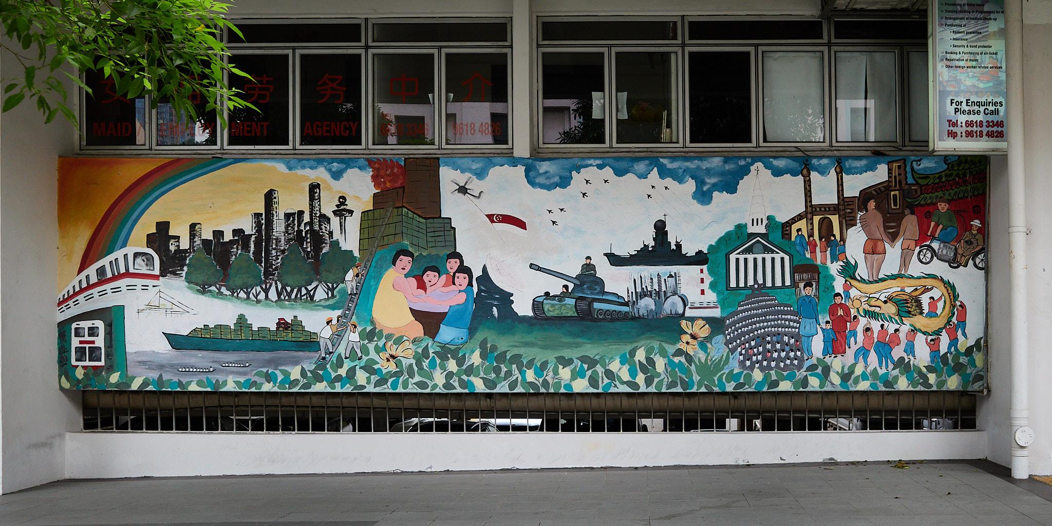 A large mural covers a wall, featuring various scenes including a train, a cargo ship, and a tank. People are depicted in different activities, with a rainbow arching above. The background includes buildings, trees, and a flag. There are also windows above the mural with red text, possibly indicating a business.