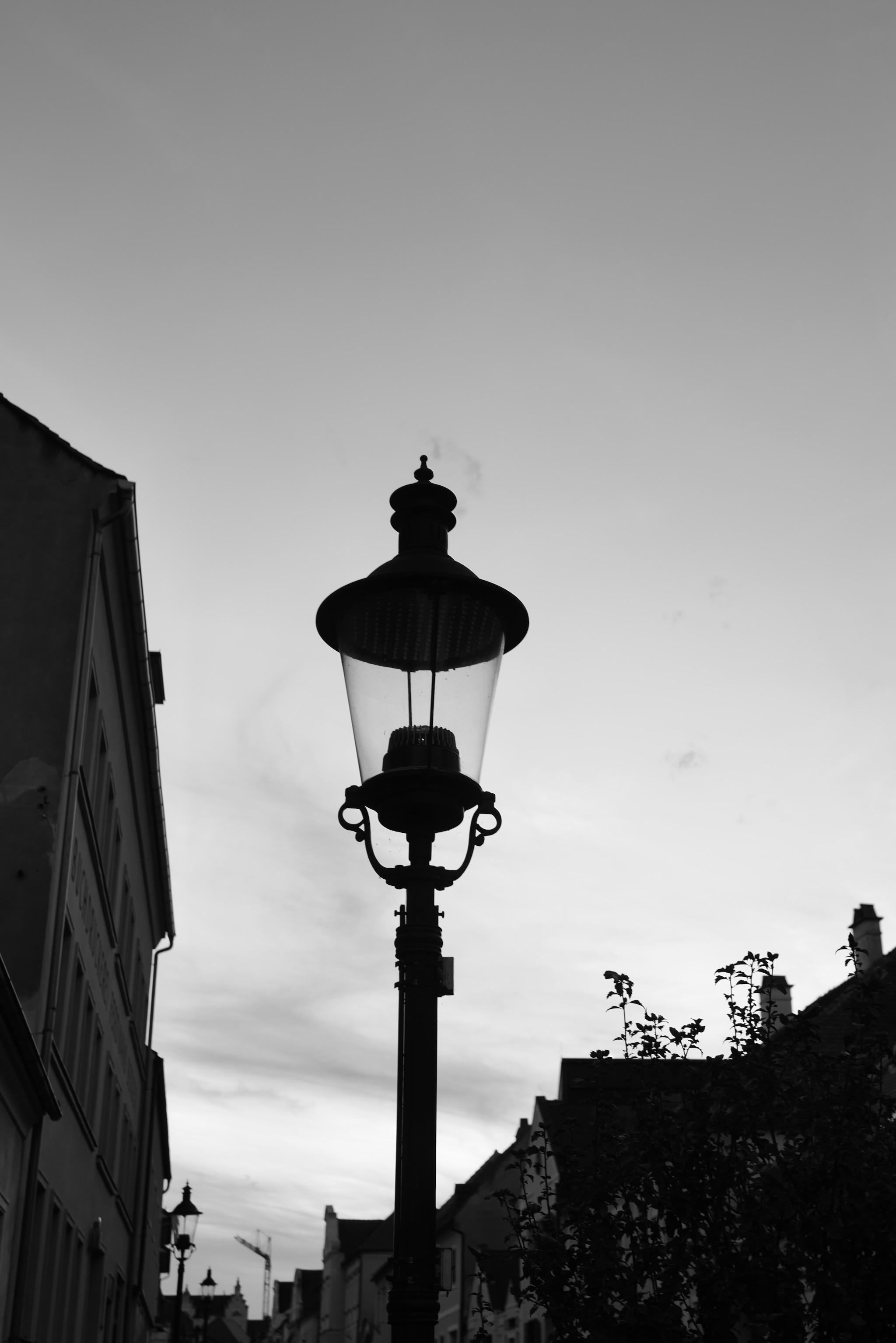 Picture of a lantern. The photo is taken from the ground against the evening sky and appears as a dark shaped silhouette.
On the left side is a building the frame, on the bottom some roofs of houses on the other side of the street. Some plants are also visible.
Picture is black and white.