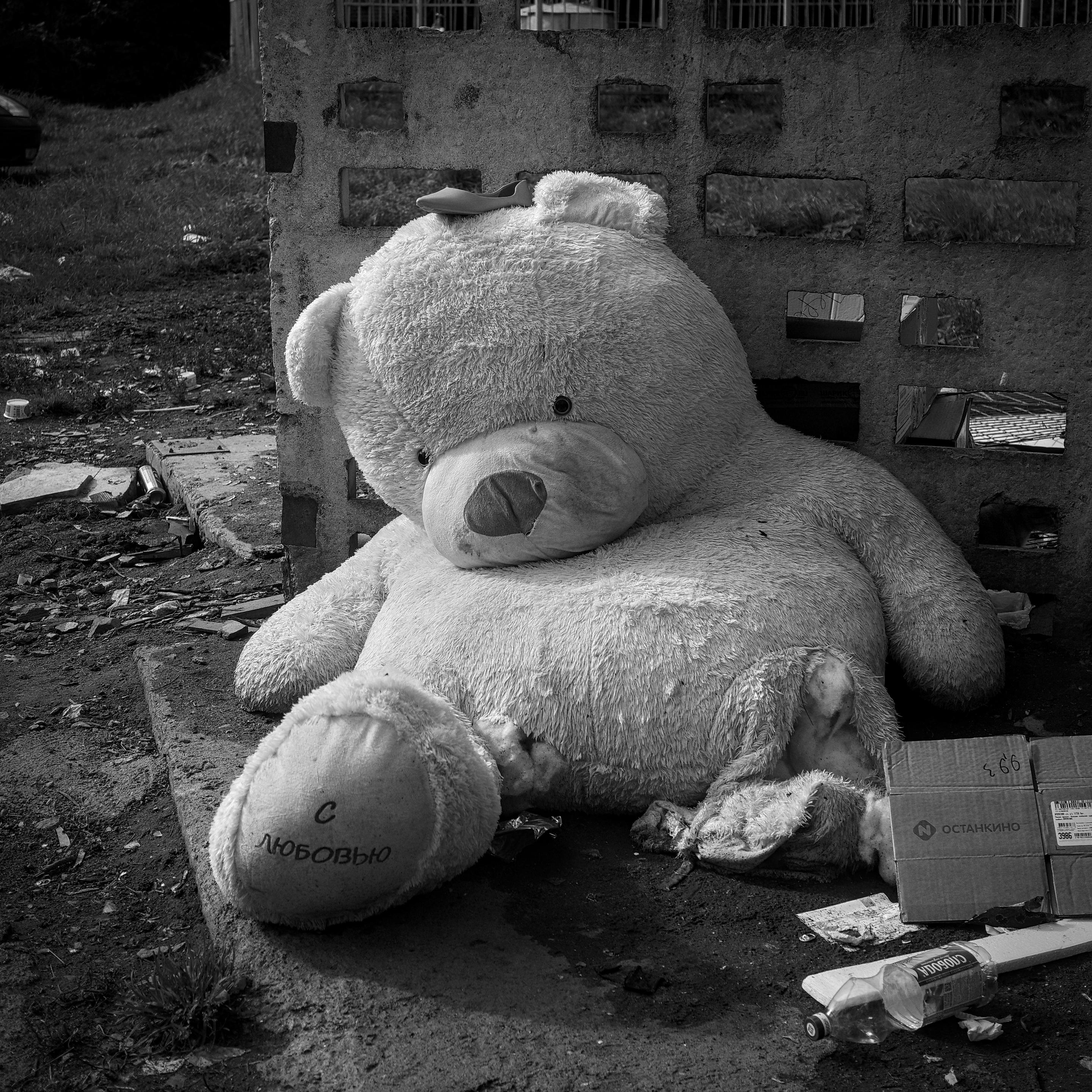 This black-and-white photograph captures a huge, discarded teddy bear sitting sadly on the ground against an old brick wall. The toy looks worn out and dirty, with its left leg torn open and its head tilted mournfully to one side. The central detail of the frame is the touching inscription "With love" ("С любовью") on the sole of its right foot. The stark contrast between this warm message and the bleak surroundings — scattered garbage, an empty bottle, and a cardboard box — creates a profound atmosphere of loneliness, abandonment, and unrequited feelings.