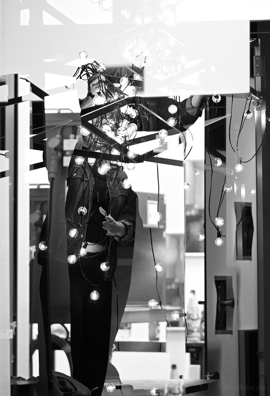 A black-and-white photo of a person standing amid tangled strings of glowing lights and geometric reflective panels in an indoor space.