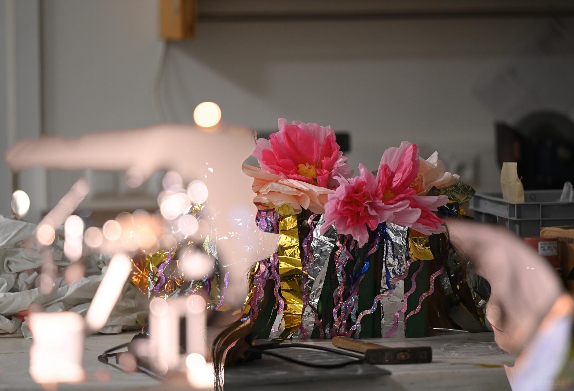 A close-up of a craft workspace featuring large pink artificial flowers arranged on a shiny gold‑and‑silver base, surrounded by ribbons, tools, and studio materials with soft bokeh reflections in the foreground.