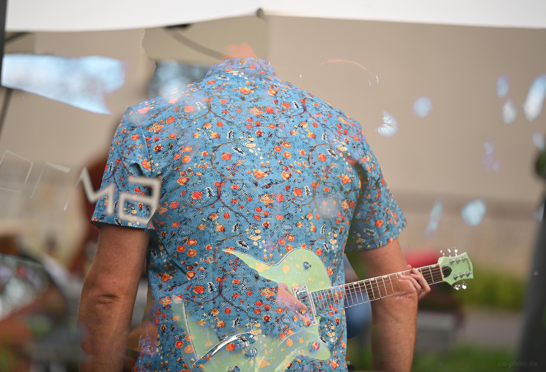 A person in a bright floral shirt plays a pastel green electric guitar, appears as a double exposure.