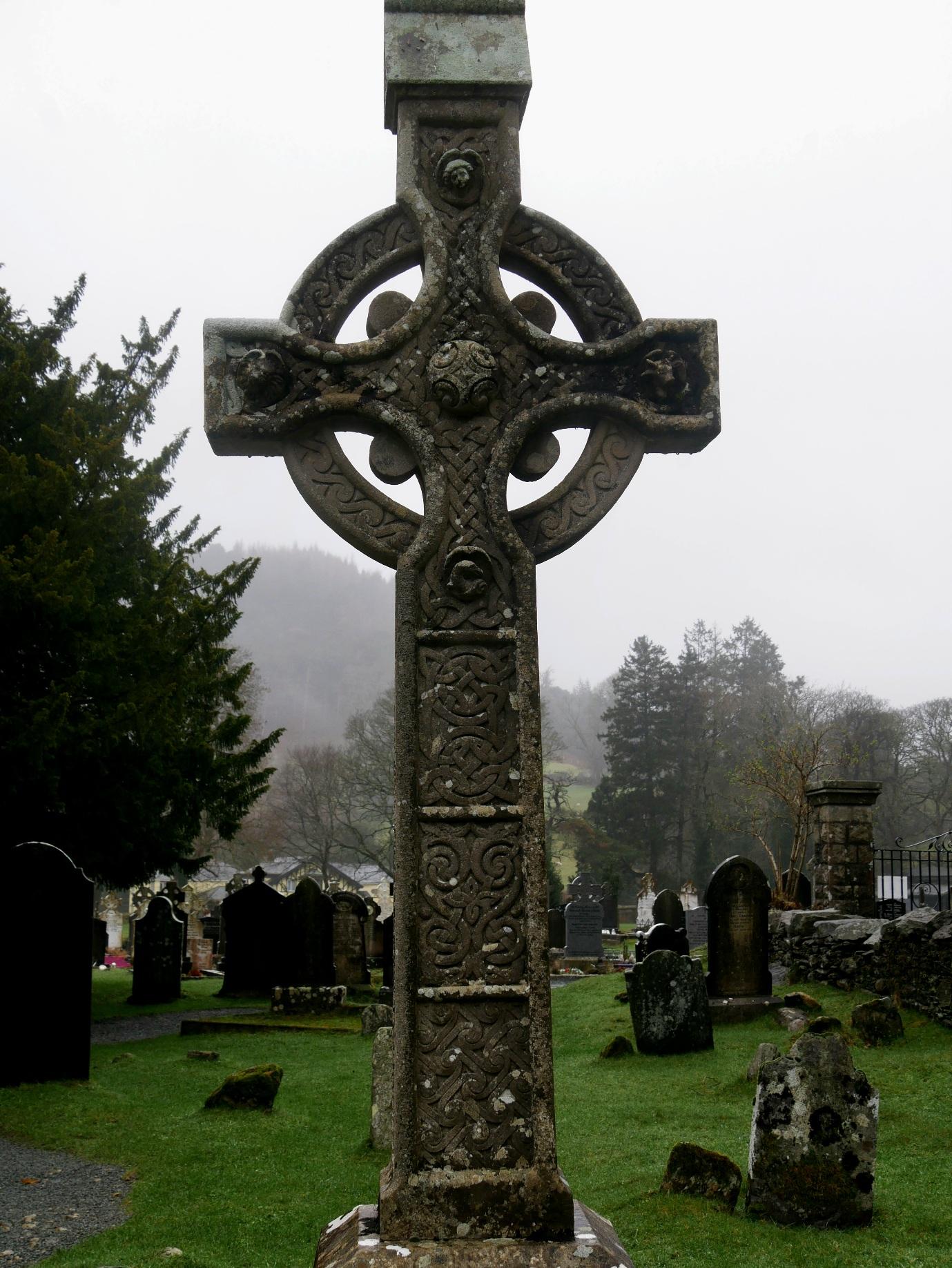 Ein altes verwittertes Hochkreuz steht auf dem Friedhof der Klosterruine Glendalough in Irland. Es ist mit spiralförmigen Ornamenten verziert. Die Stimmung ist aufgrund des nebligen Wetters mystisch. Irland 2024