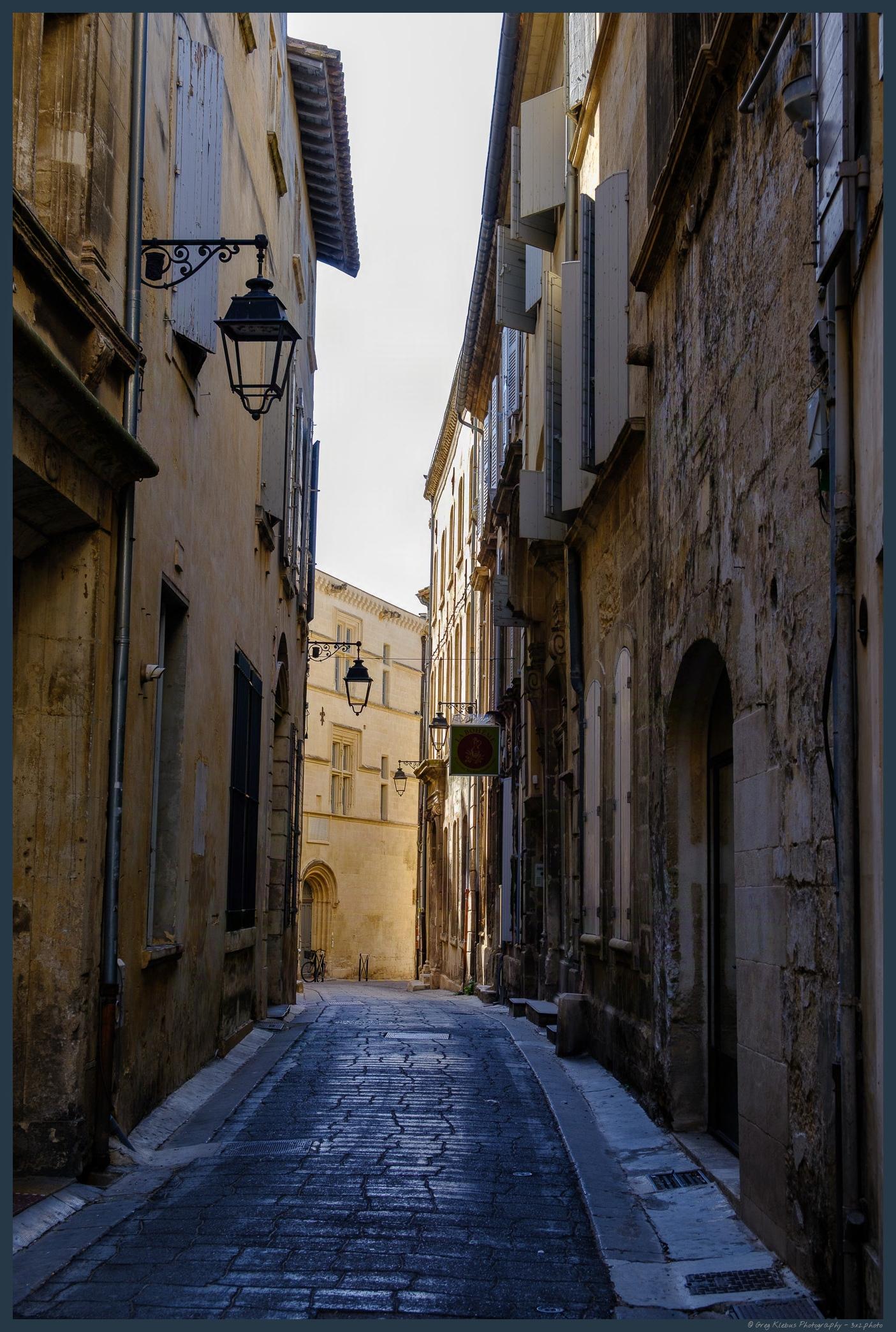 Narrow, shadowed cobblestone alley lined with weathered stone and stucco buildings, hanging lanterns, balconies, and a sunlit street in the distance.
