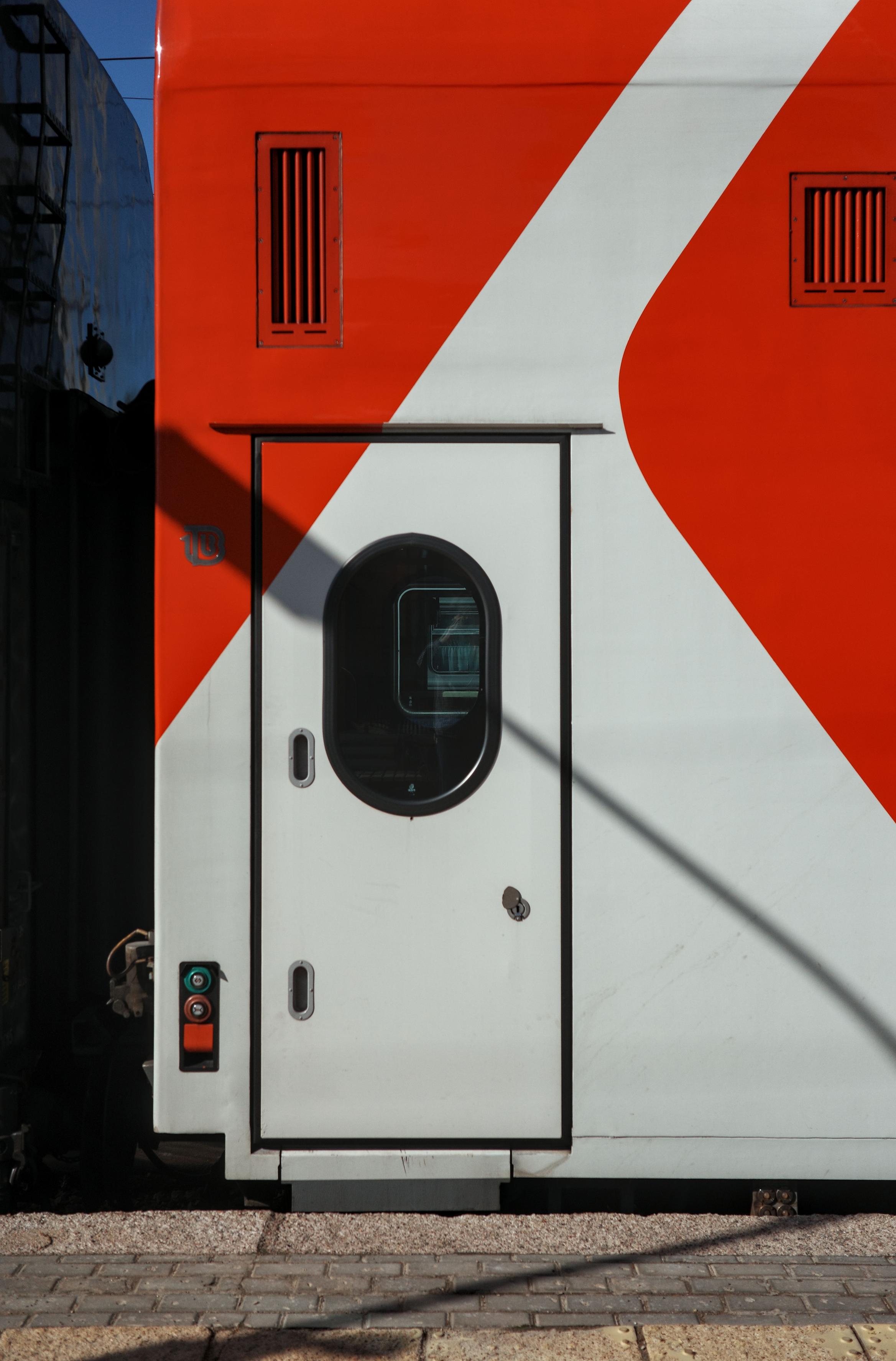 A close-up of a train car door with a bright red-and-white geometric pattern. The oval window, ventilation grilles, and the clear line of shadow create a powerful architectural composition.