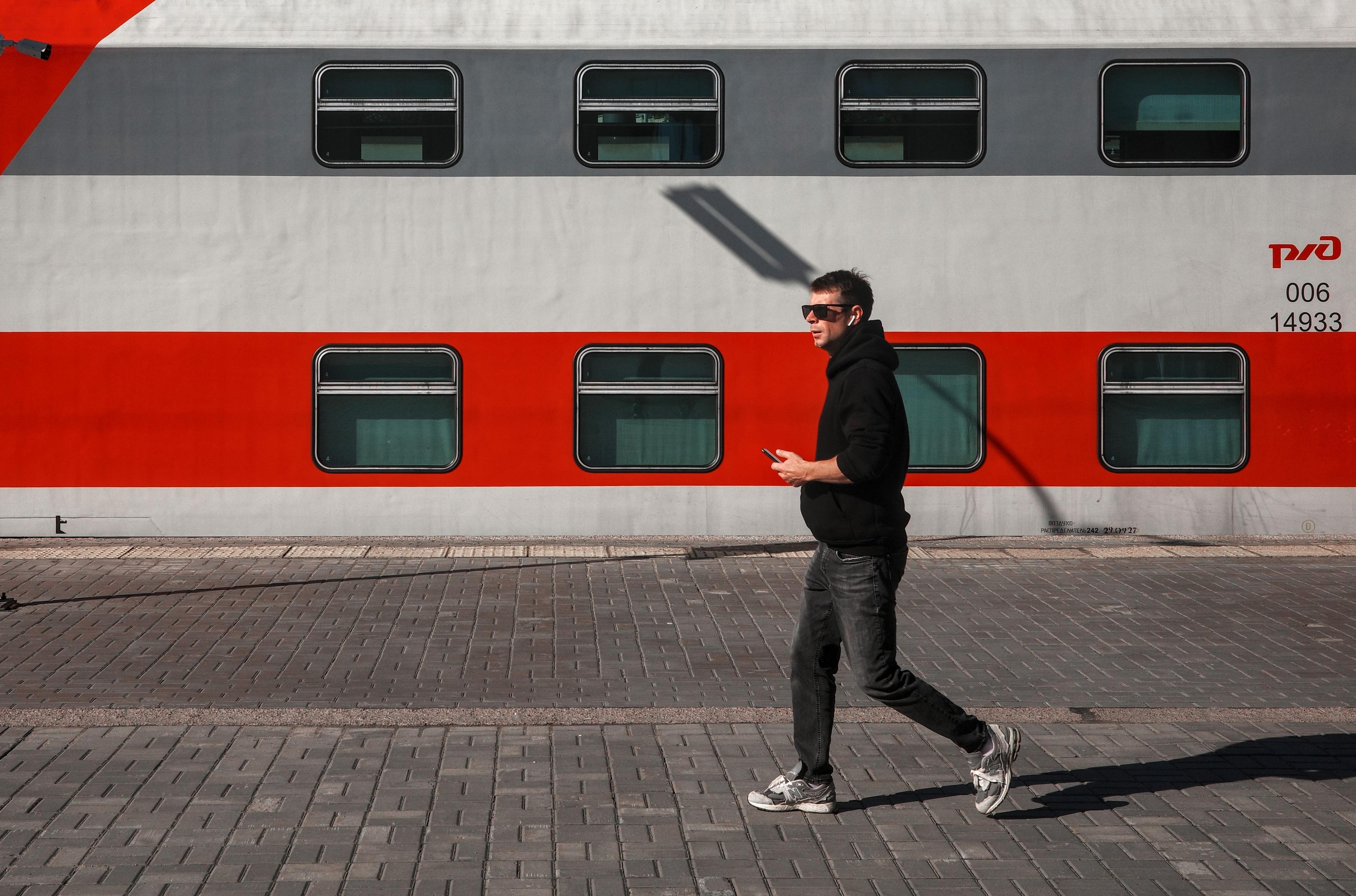 A young man in dark clothes and sunglasses walks along the platform alongside the train. His movement and shadow create dynamism against the static red car. The image conveys the atmosphere of a lively urban life.