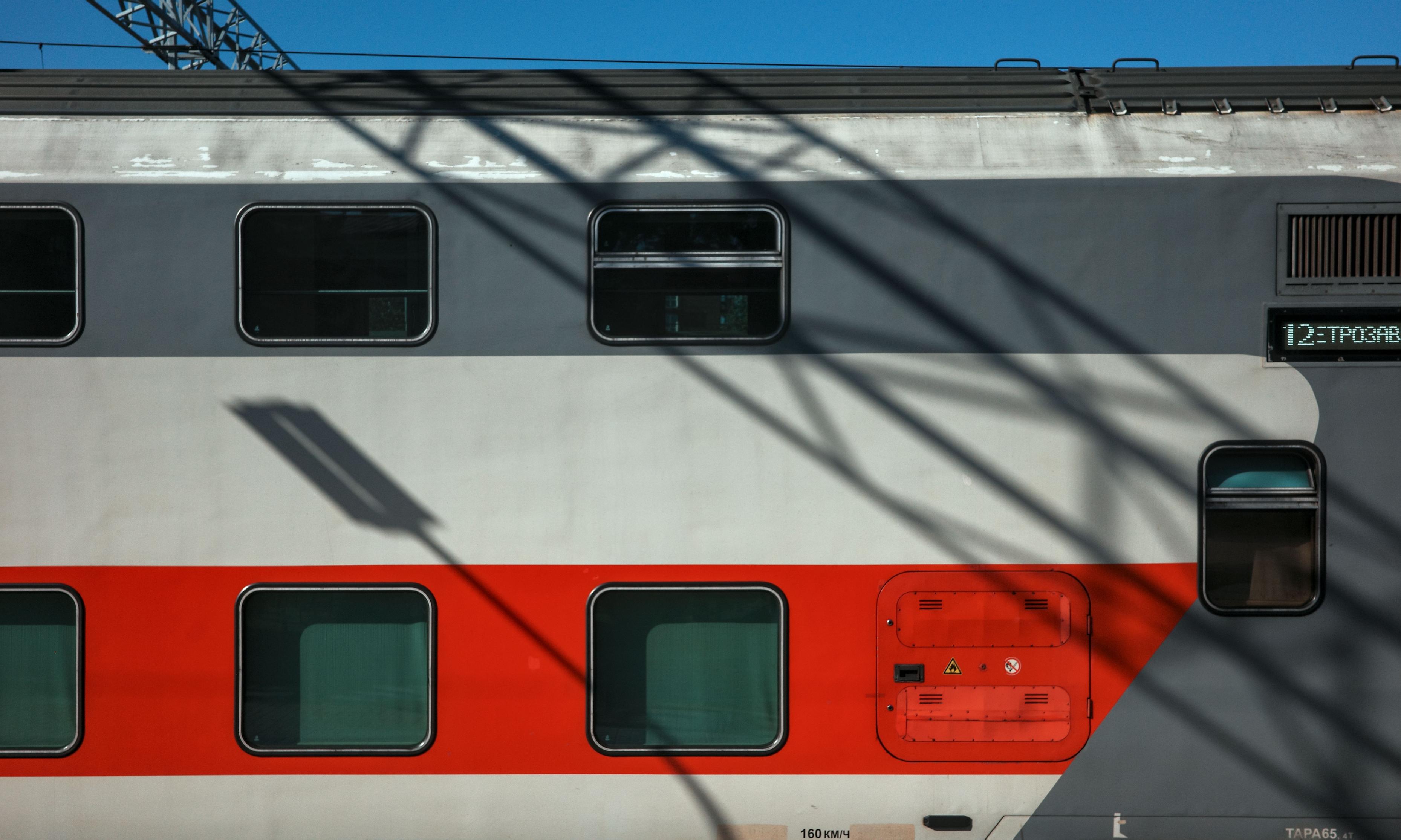 A close-up of the side of a train car. Shadows from railway structures falling on the red-and-white body create a complex, almost abstract graphic pattern. The image highlights the interplay of light and shadow on the train's surface.