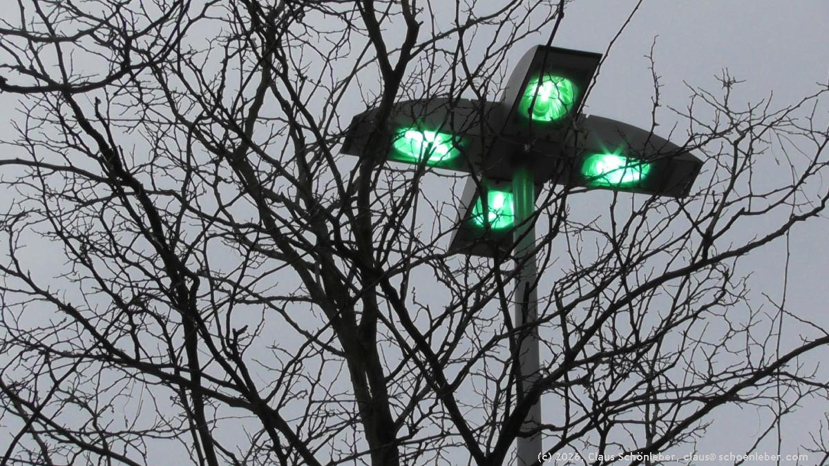Im Vordergrund die Krone eines Baumes ohne Blätter (Winter) , aufgenommen von unten, einige Meter entfernt.

Dahinter eine sehr große Straßenlaterne, die einen ganzen Platz beleuchtet.
Sie hat kreuzförmig vier Ausläfer, die jeweils eine sehr helle Gasdrucklampe enthalten. Die Lampen sind allerdings gerade eben erst eingeschaltet worden (Dämmerungsschaltung) und heizen auf, daher leuchten sie noch recht dunkel in einem Türkisgrün. Die Lampe ist ungefähr sieben Meter hoch.

Der Himmel ist grau.
