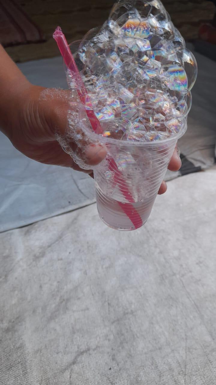 A hand holds a clear plastic cup filled with water and a pink straw, creating a large, overflowing mound of iridescent bubbles.