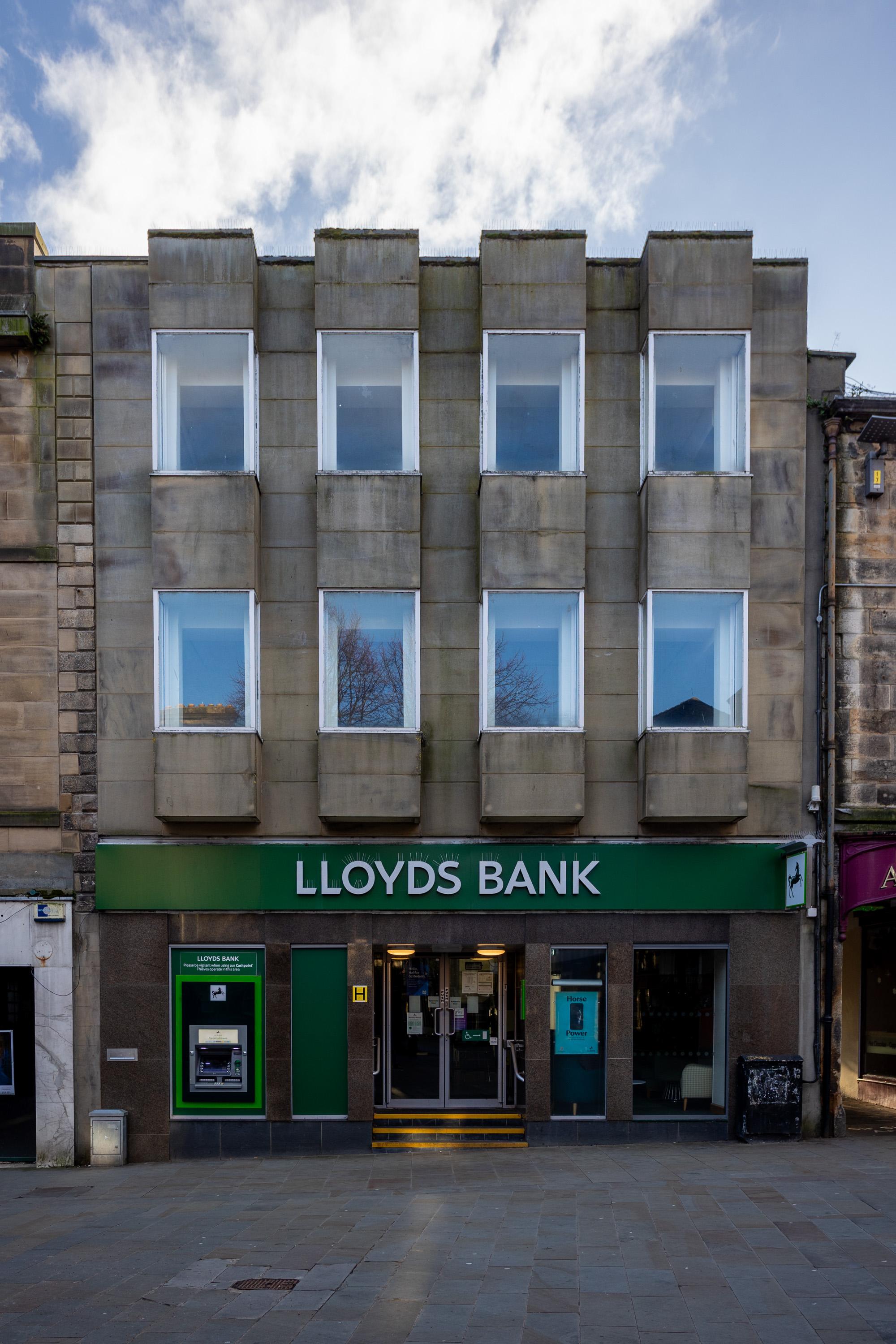 A Lloyds Bank branch with a modern stone façade and six white-framed windows above a green sign. Two cashpoints and a glass entrance door are at street level. The pavement is empty, and the sky is partly cloudy.