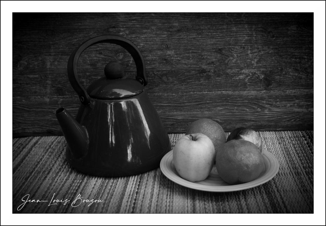 This is a black and white still life image featuring a  teapot and a plate of fruit against a rustic wooden background.

The main elements are a large, dark  teapot positioned on the left side of the frame, and a small, light-colored plate containing several pieces of fruit on the right. The teapot is round and bulbous with a prominent black handle arching over the top, and a small, rounded lid knob. Its spout juts out to the left. The surface of the teapot is highly reflective, showing bright highlights that accentuate its curved form.

The plate of fruit is placed in front of the teapot, slightly to the right. It holds at least three fruits: a pale yellow-green apple is prominent in the foreground, to the left of the plate's center. Behind it and slightly to the right is a darker, round fruit, possibly an orange or a plum, with some texture visible. To the right of the apple and slightly further back is another round, darker fruit, appearing similar to the one behind the apple. The p