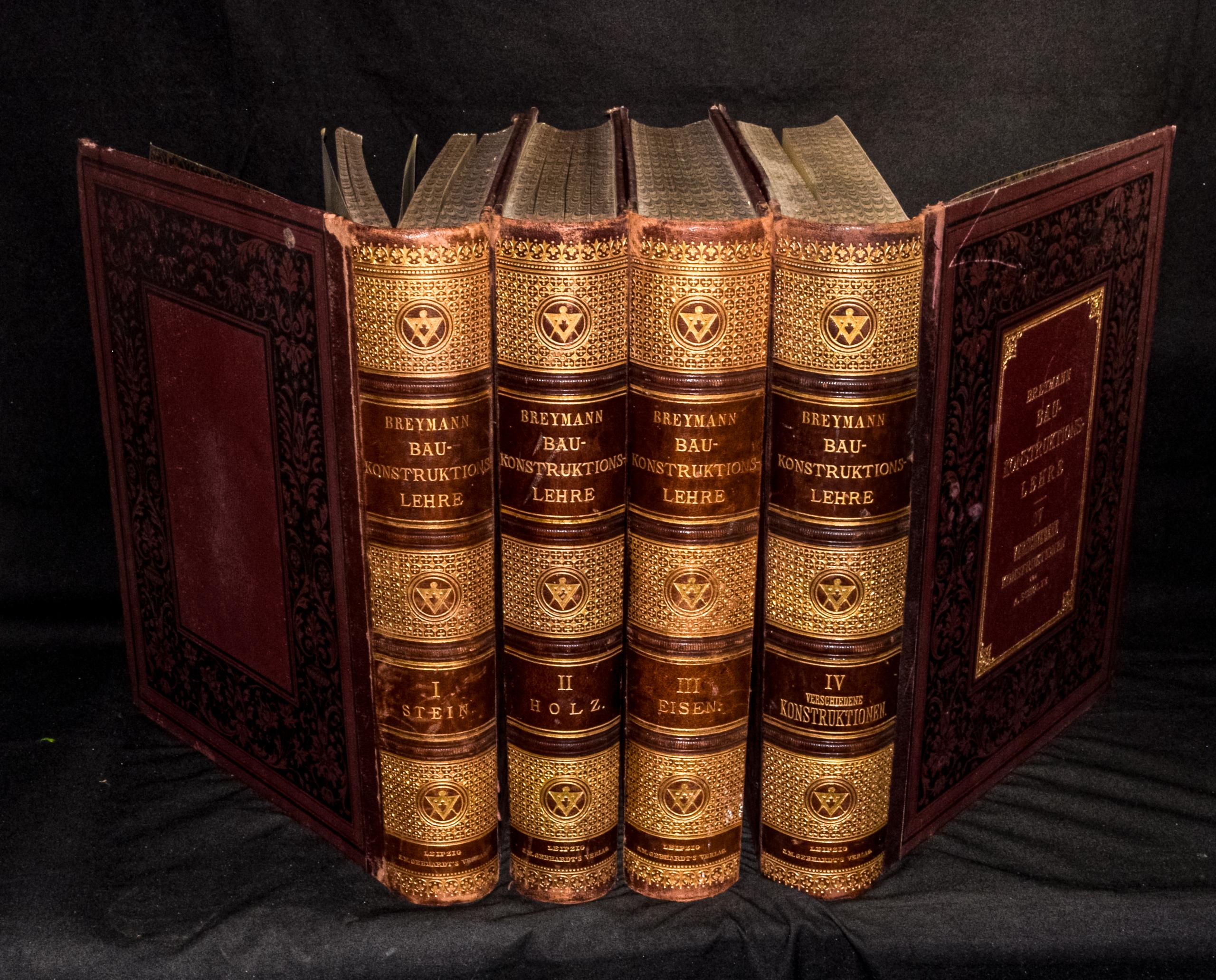 Vierteiliges Set historischer, reich verzierter Prachtbände der „Breymann Bau-Konstruktions-Lehre“, aufgefächert vor schwarzem Hintergrund aufgestellt. Die braunen Lederrücken weisen aufwendige Goldprägungen mit geometrischen Ornamenten, den Bandtiteln sowie den Bandbezeichnungen „I. Stein“, „II. Holz“, „III. Eisen“ und „IV. Verschiedene Konstruktionen“ auf. Die beiden äußeren Bände sind nach außen geöffnet, sodass die aufwendig gemusterten Innenseiten und der vergoldete Deckeltitel sichtbar werden.