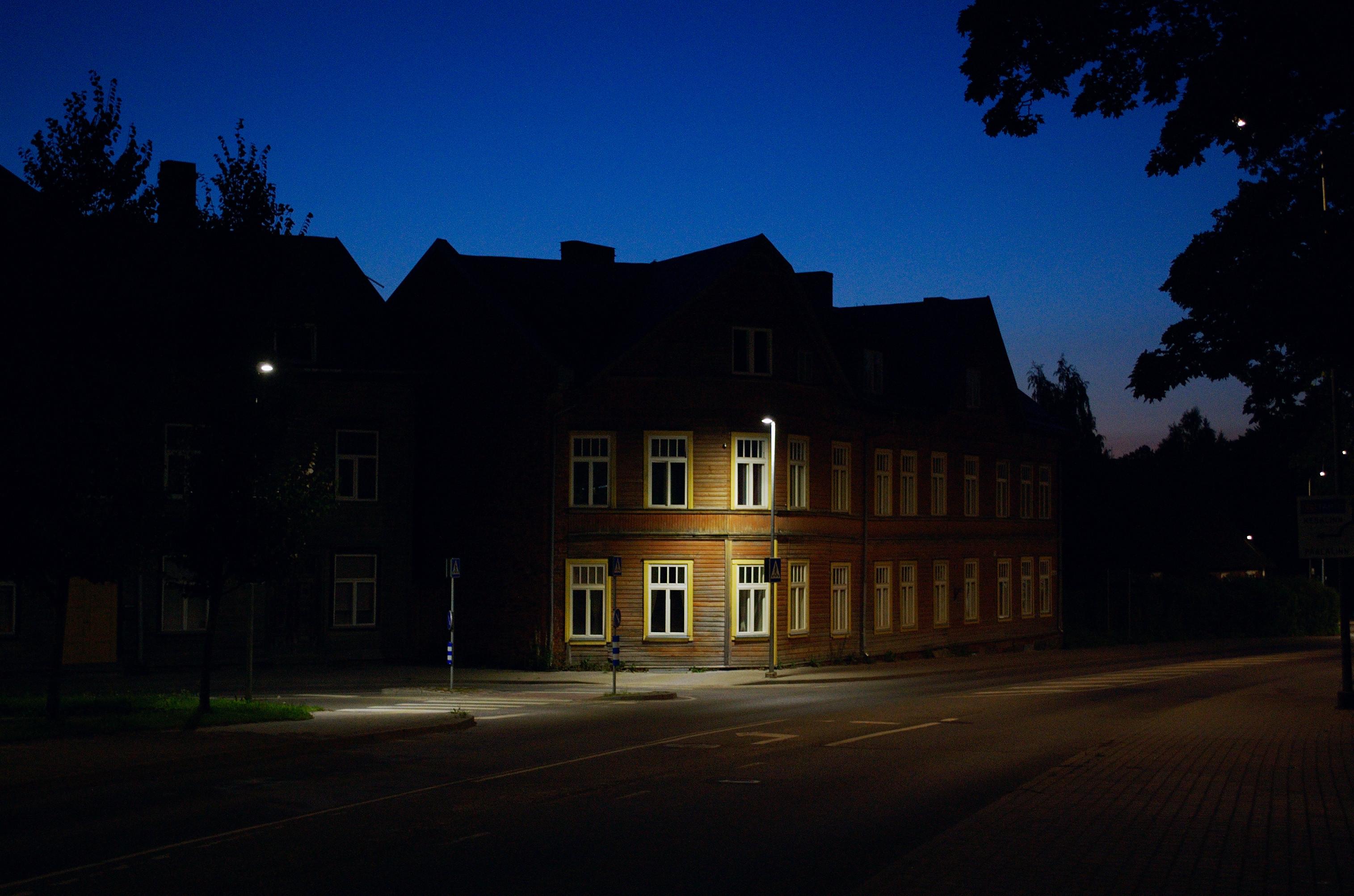 Photo of a brown wooden two-storey house at night. Most of the house and the trees at the edges of the frame are only visible as black silhouettes, stark against a deep blue sky. A single streetlight picks out a part of the house’s facade and casts a patch of light on the ground in front of it.