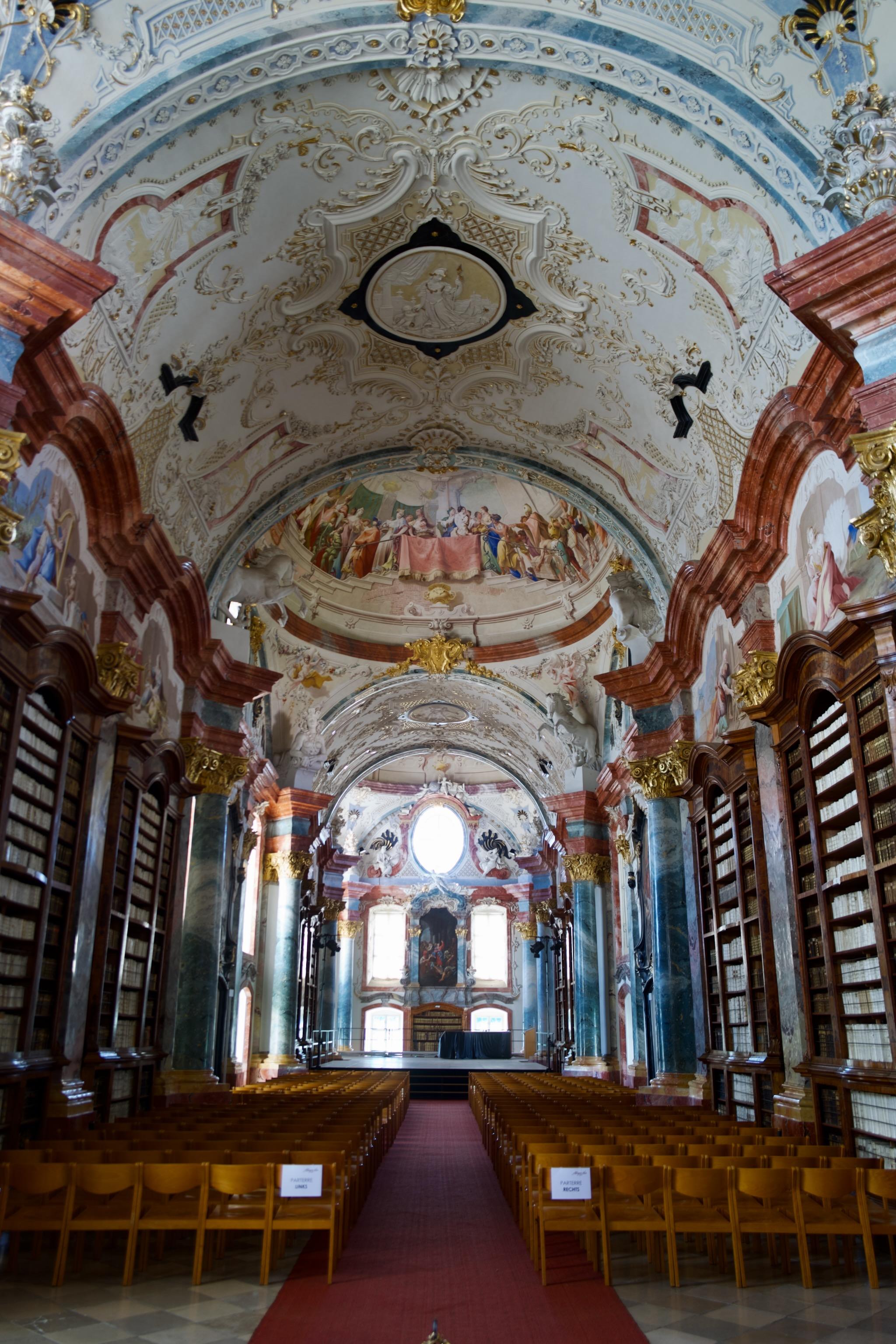 Picture of a baroque library in a cloister in Austria. It is heavily ornamented and painted. Main colors are brown fake marble, white stucco and bright blue applications.
Left and right are books shelfs up to several meters. On the floor are chairs prepared for an event or concert.