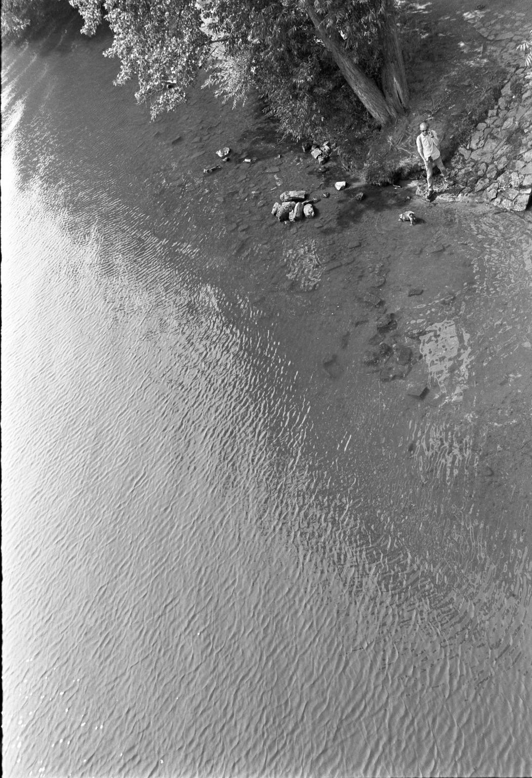 A black and white image of a calm river with gentle ripples. There are rocks visible at the riverbank, and a person stands on the shore, partially shaded by a tree. The scene conveys a tranquil, natural setting.