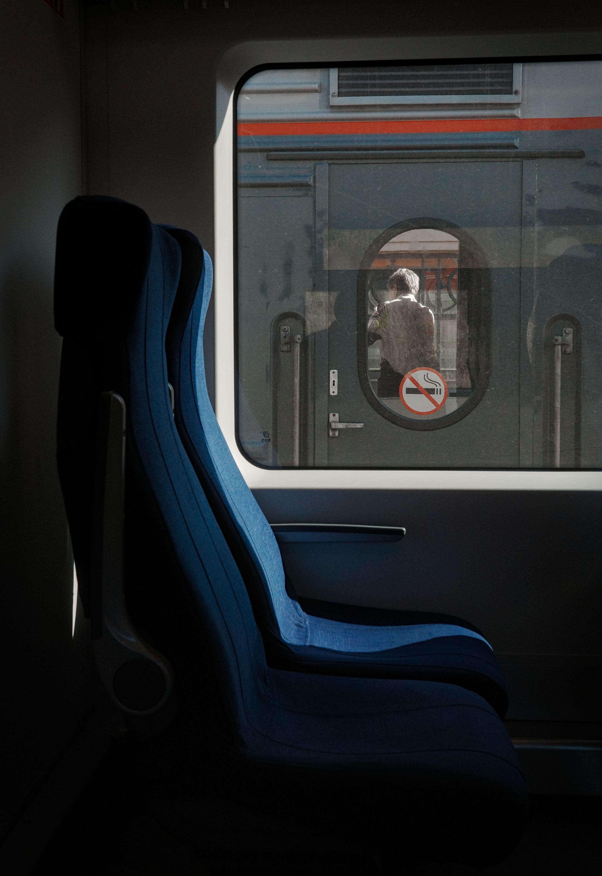 A view from inside a train carriage. The dark blue seats create a cozy but cold atmosphere. Another train and the silhouette of a person are reflected in the window, and a "No Smoking" sign is visible. The image conveys a sense of travel and looking at the world from within.