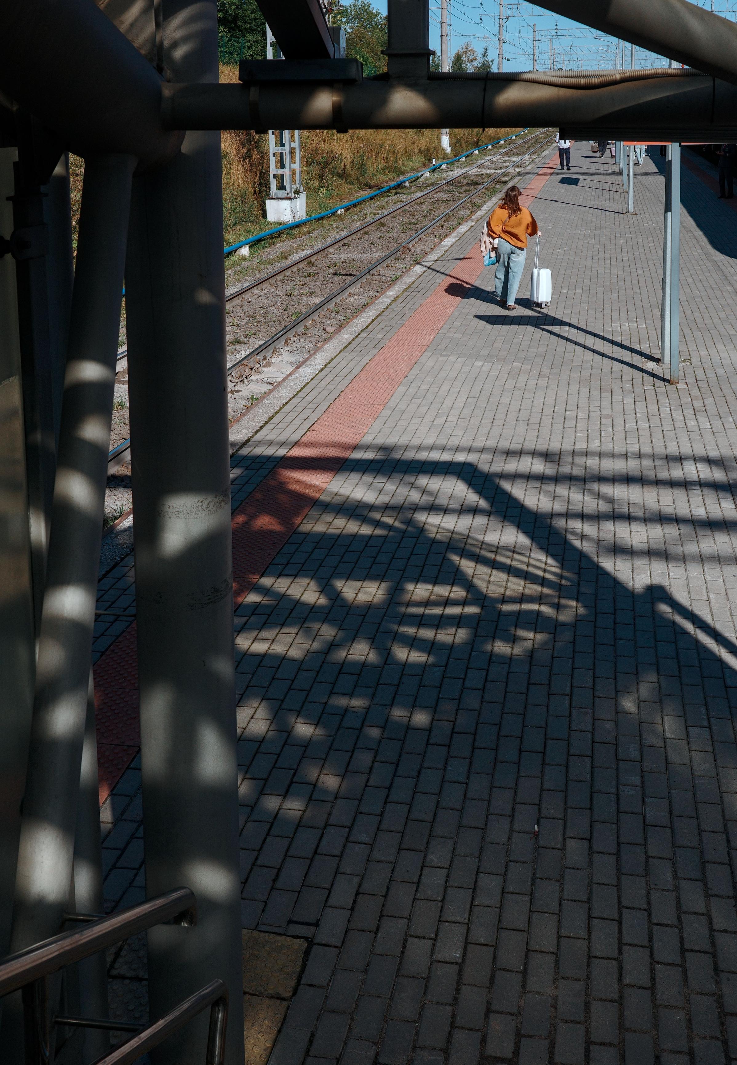 A wide shot from the platform. Shadows from the canopy fall on the tiles, creating a graphic pattern. A woman in an orange jacket with a suitcase walks toward the camera, her figure setting the scale and direction of movement.